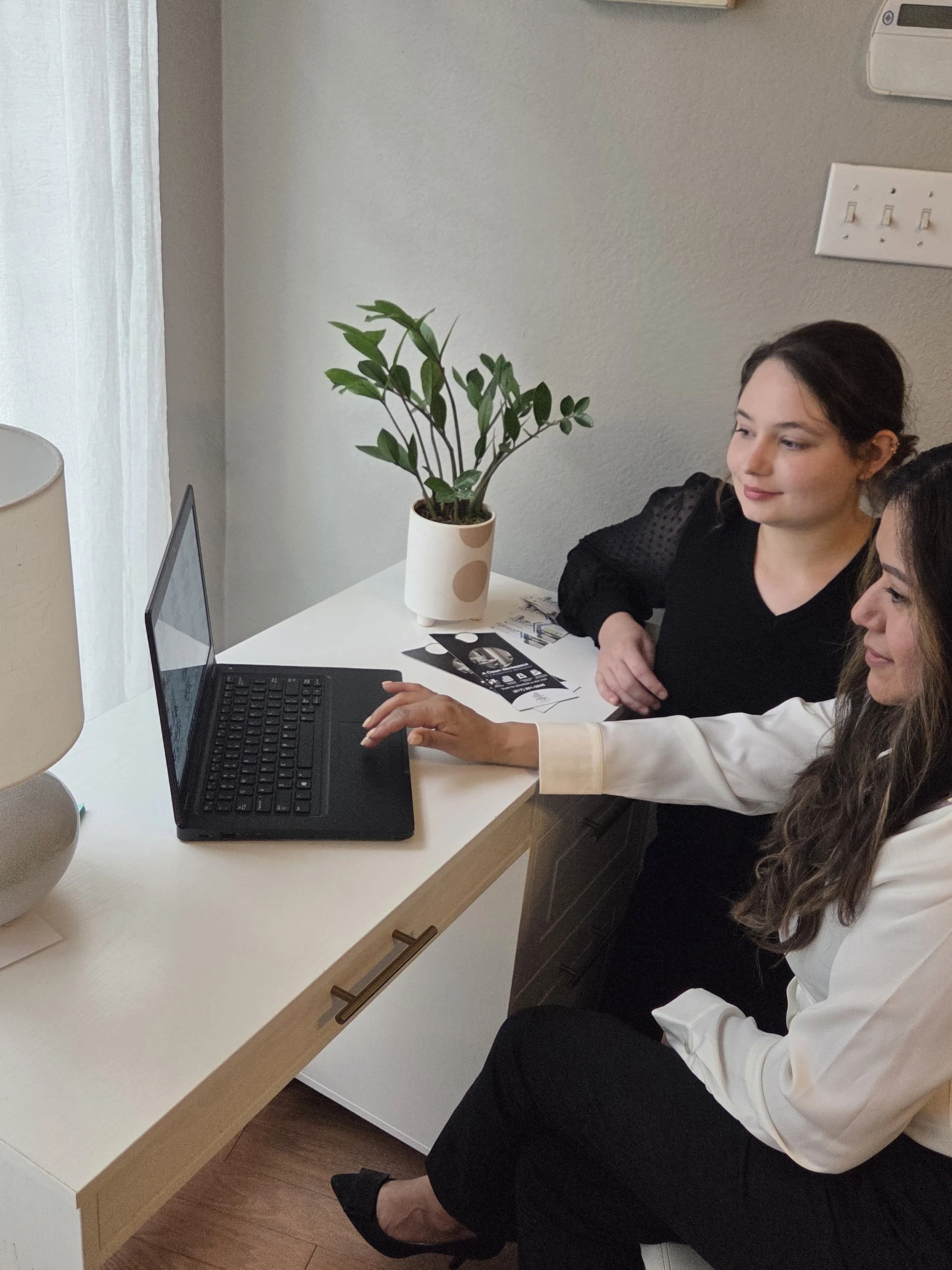 Two women sitting at a white desk looking at a laptop screen. One woman is pointing at the screen, and there is a potted plant and some papers on the desk.