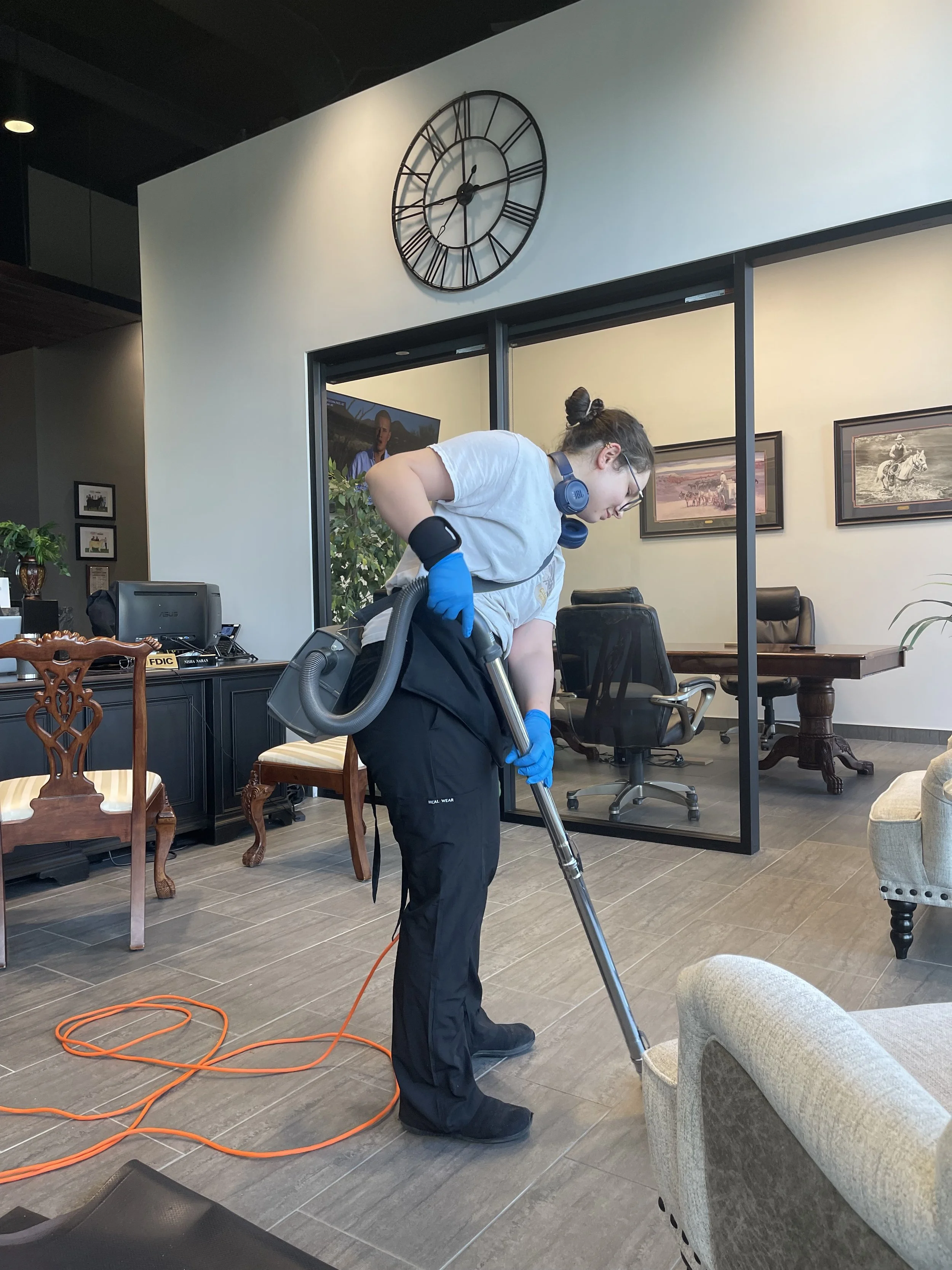 A woman wearing blue gloves, a gray t-shirt, and black pants is cleaning the floor with a vacuum cleaner in an office or conference room. She has a headset around her neck and is focused on her task.