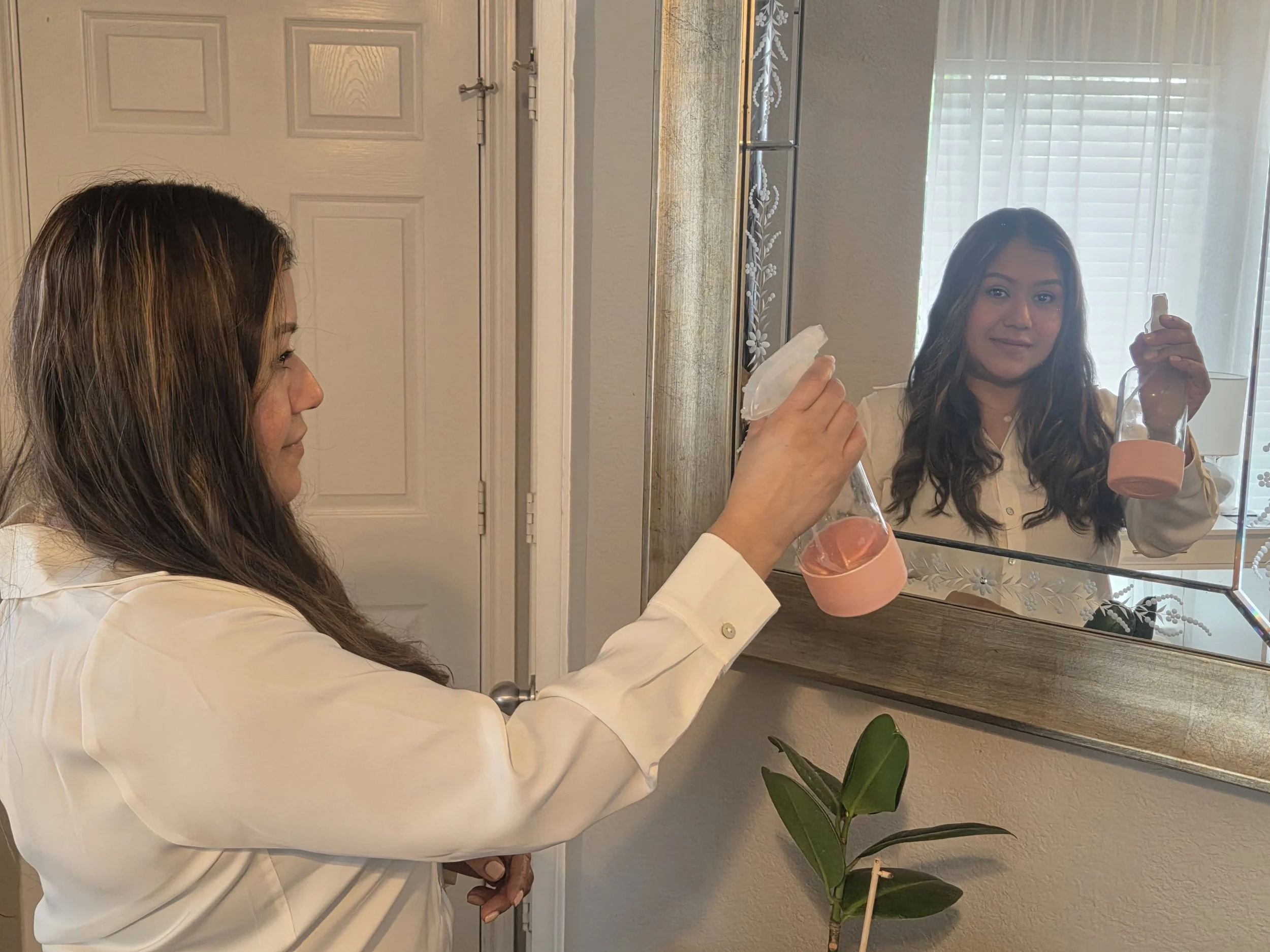 Woman in white shirt holding a pink drink and a straw, looking at herself in the mirror.