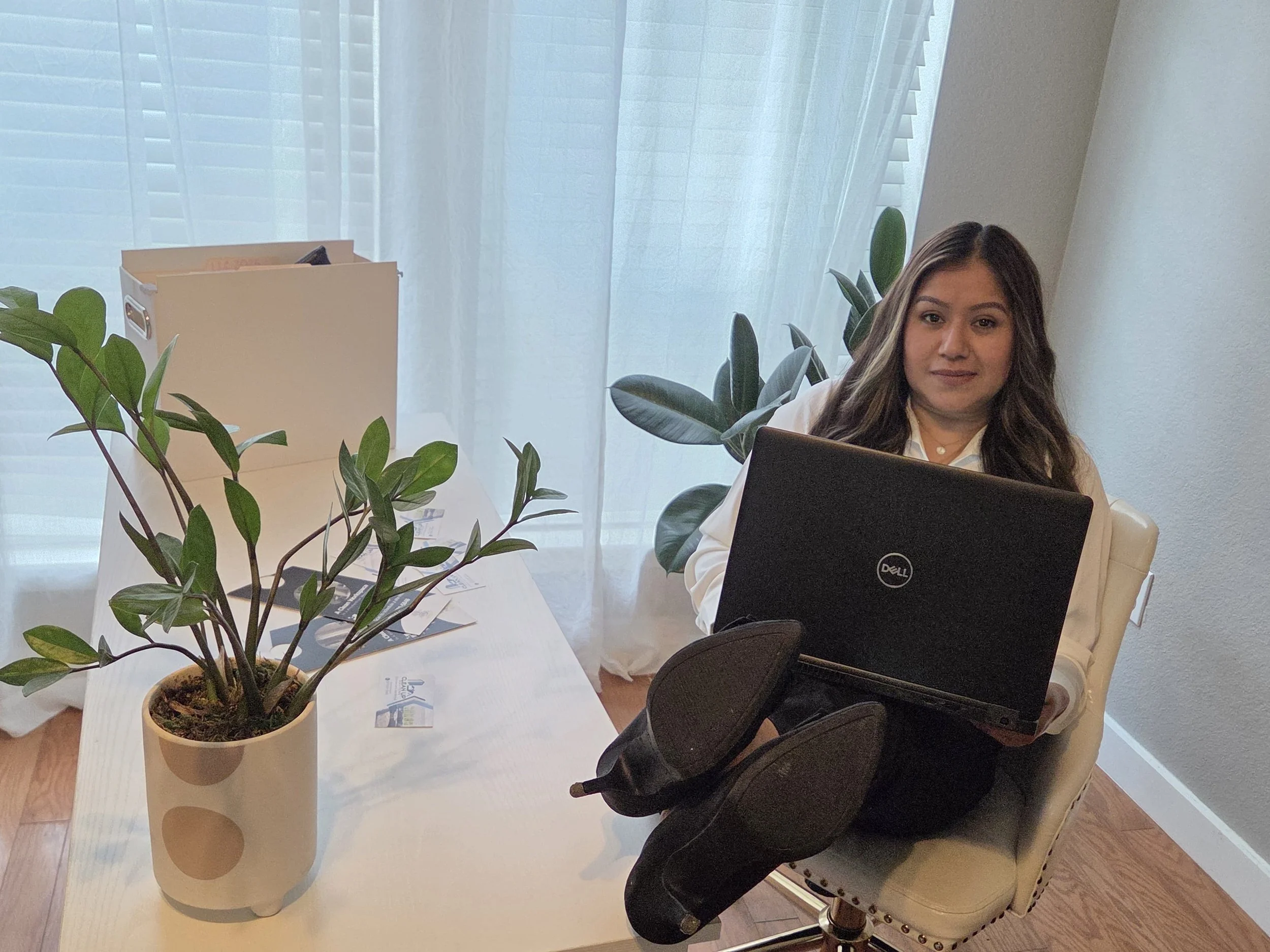 A woman sits at a white desk with a laptop on her lap. She has long dark hair and is wearing a white shirt. There are green plants, a white folder, and some scattered papers on the desk. She is sitting in a beige chair near a window with sheer white 