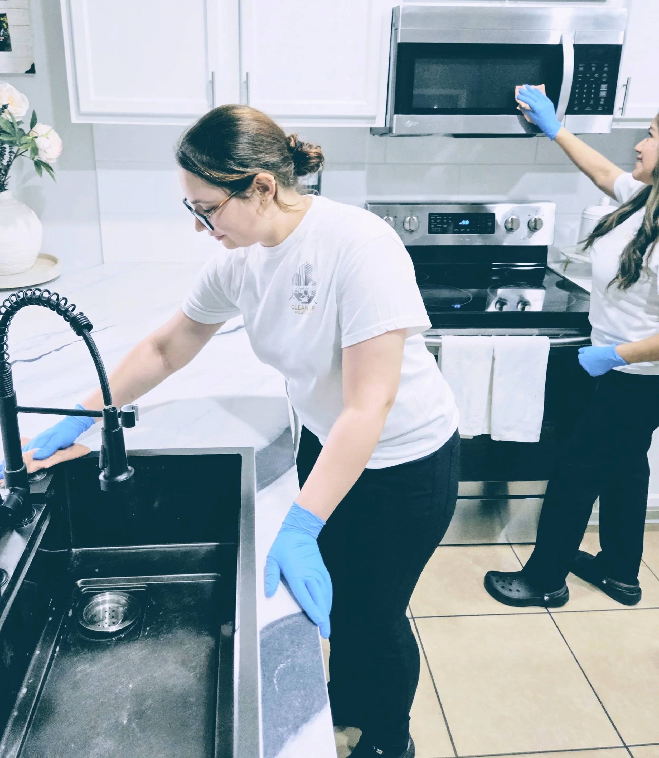 Two women in a kitchen cleaning dishes with blue gloves. One is washing dishes at the sink and the other is spraying something on a microwave.
