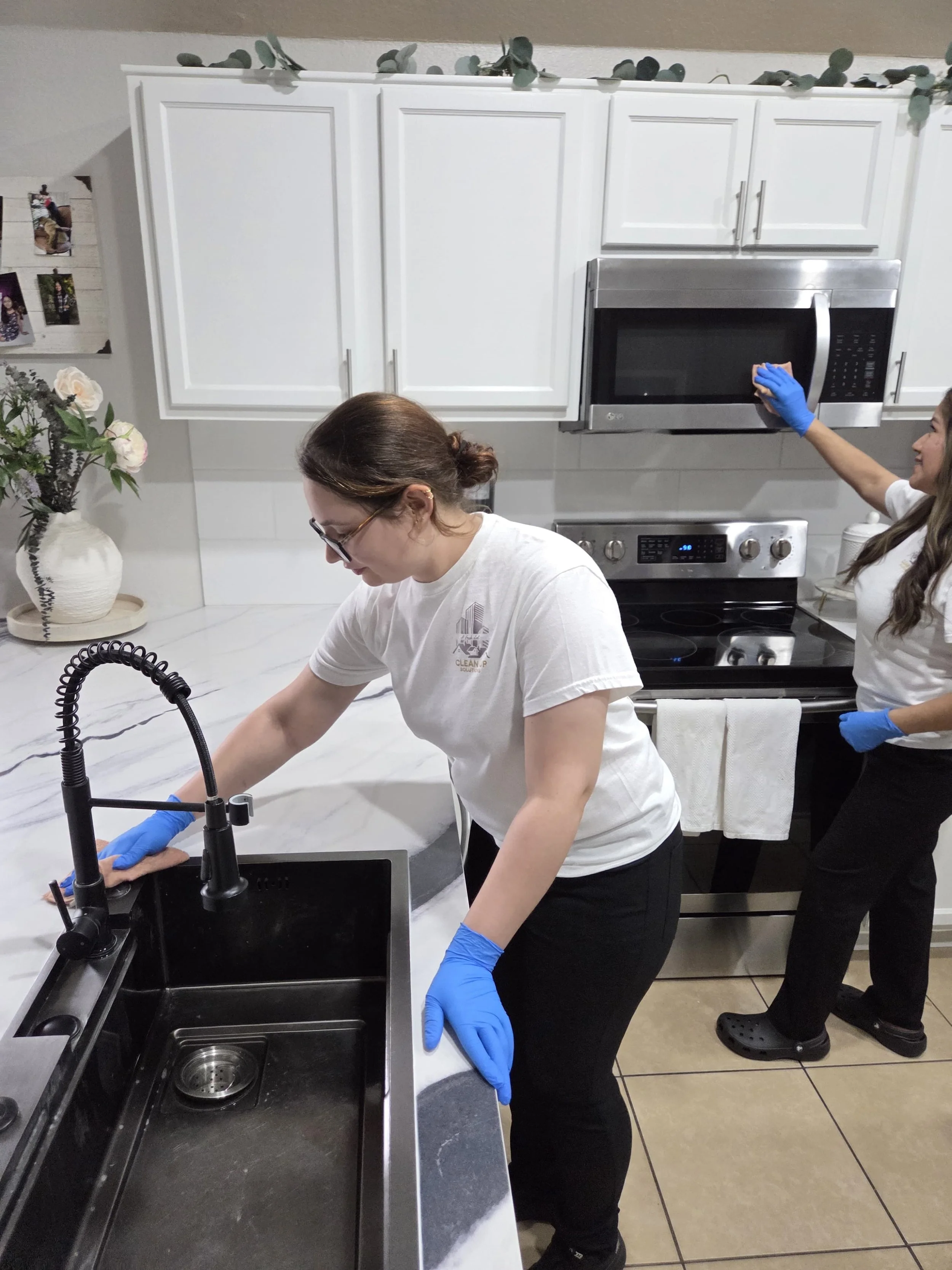 Two women cleaning a kitchen with white cabinets, a stainless steel microwave, and a stove. One woman is washing the counter near the sink, and the other is wiping the microwave door. Both are wearing blue gloves and casual clothes.