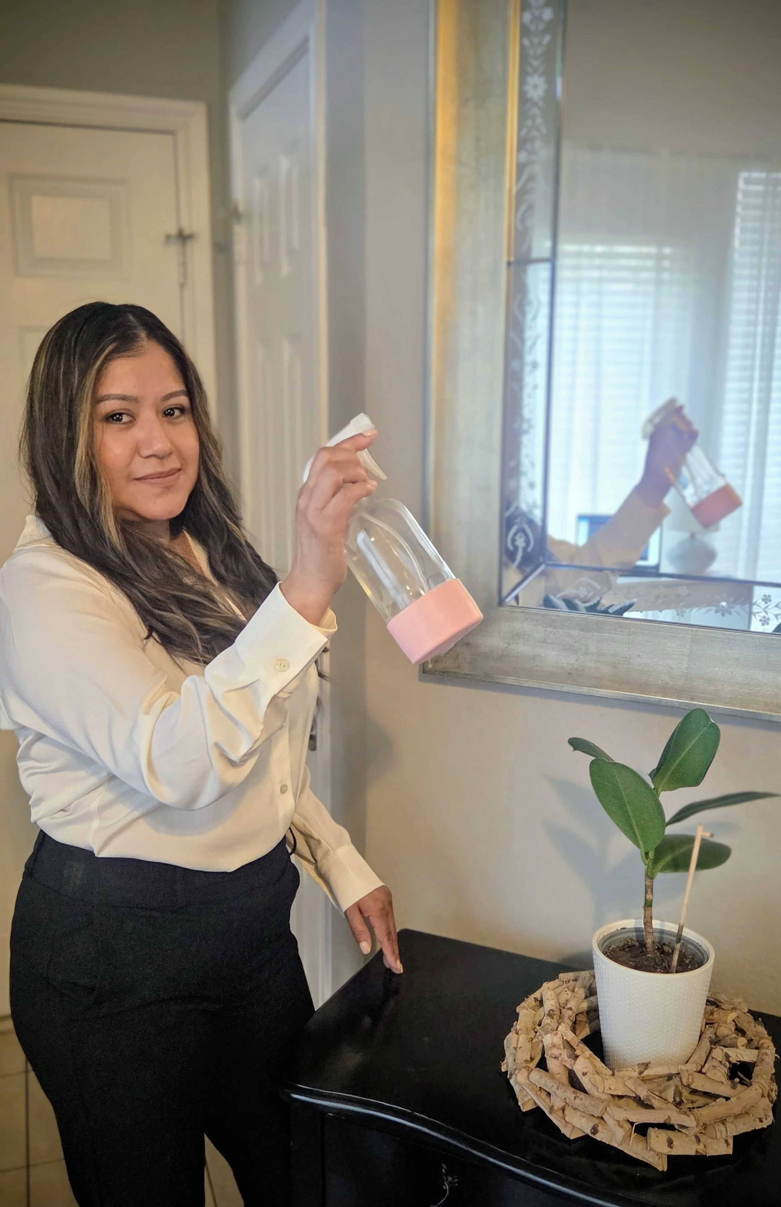 A woman in a cream-colored shirt and black pants is spraying a potted plant on a black table while standing indoors. The plant is in a white pot placed on a cardboard base. There is a mirror on the wall behind her reflecting her image and a window wi