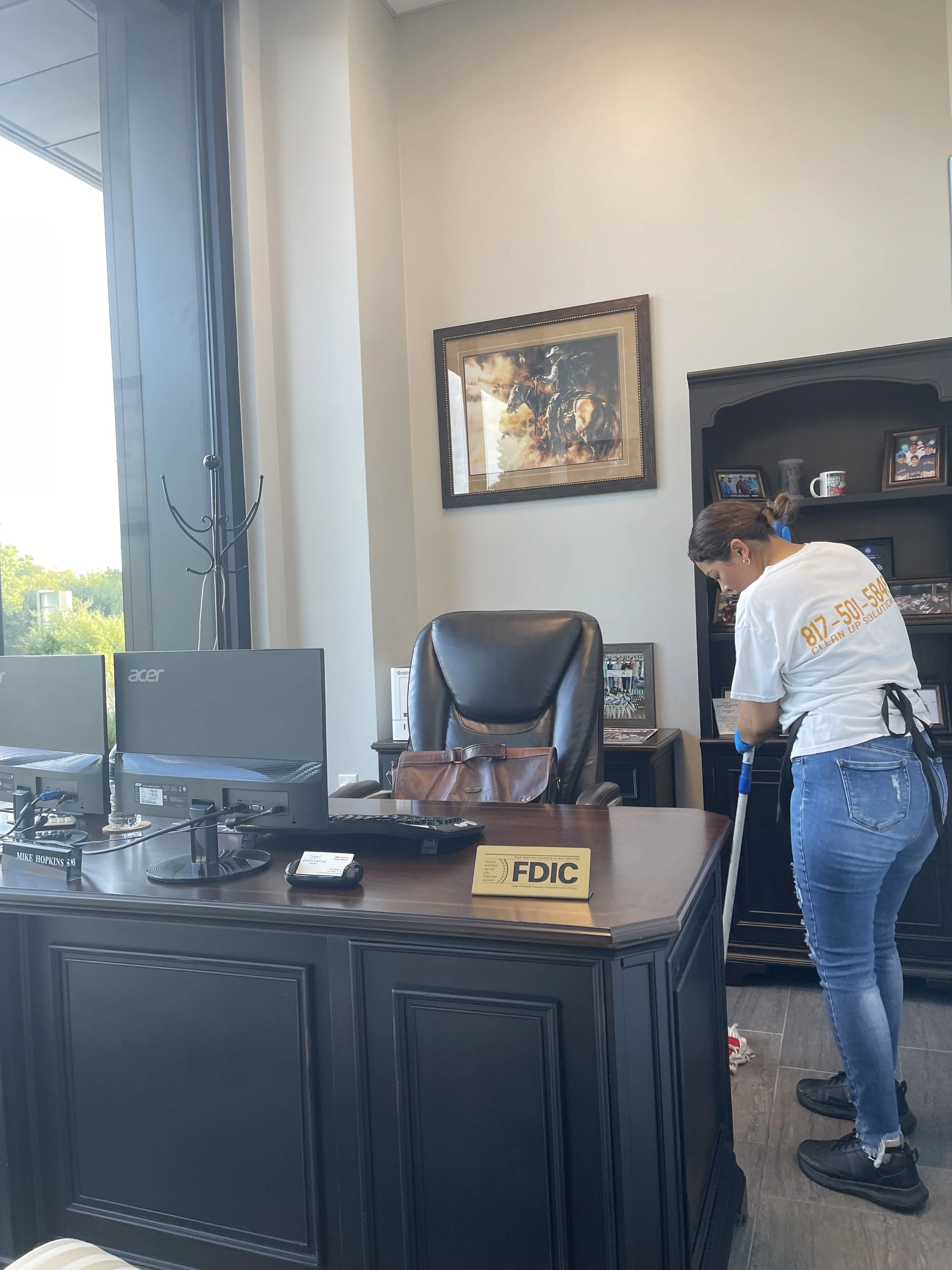 A woman cleaning an office with a mop behind a large dark wood desk. The office has a window and a painting on the wall. The desk has two computer monitors, a nameplate, and some office supplies. The woman is wearing a white T-shirt, jeans, and black