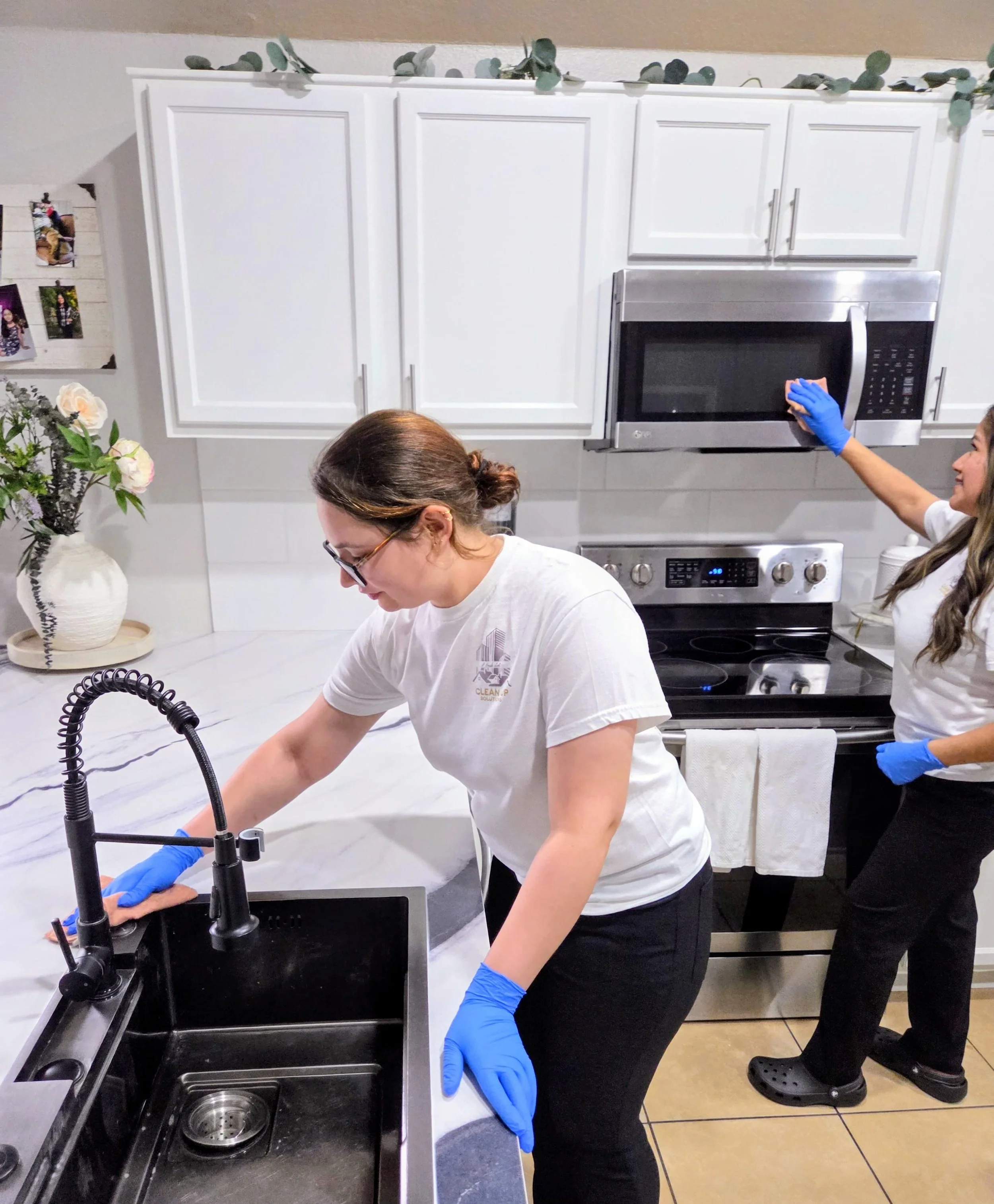 Two women cleaning a modern kitchen; one scrubbing the sink and the other wiping the microwave, both wearing blue gloves and casual white shirts.