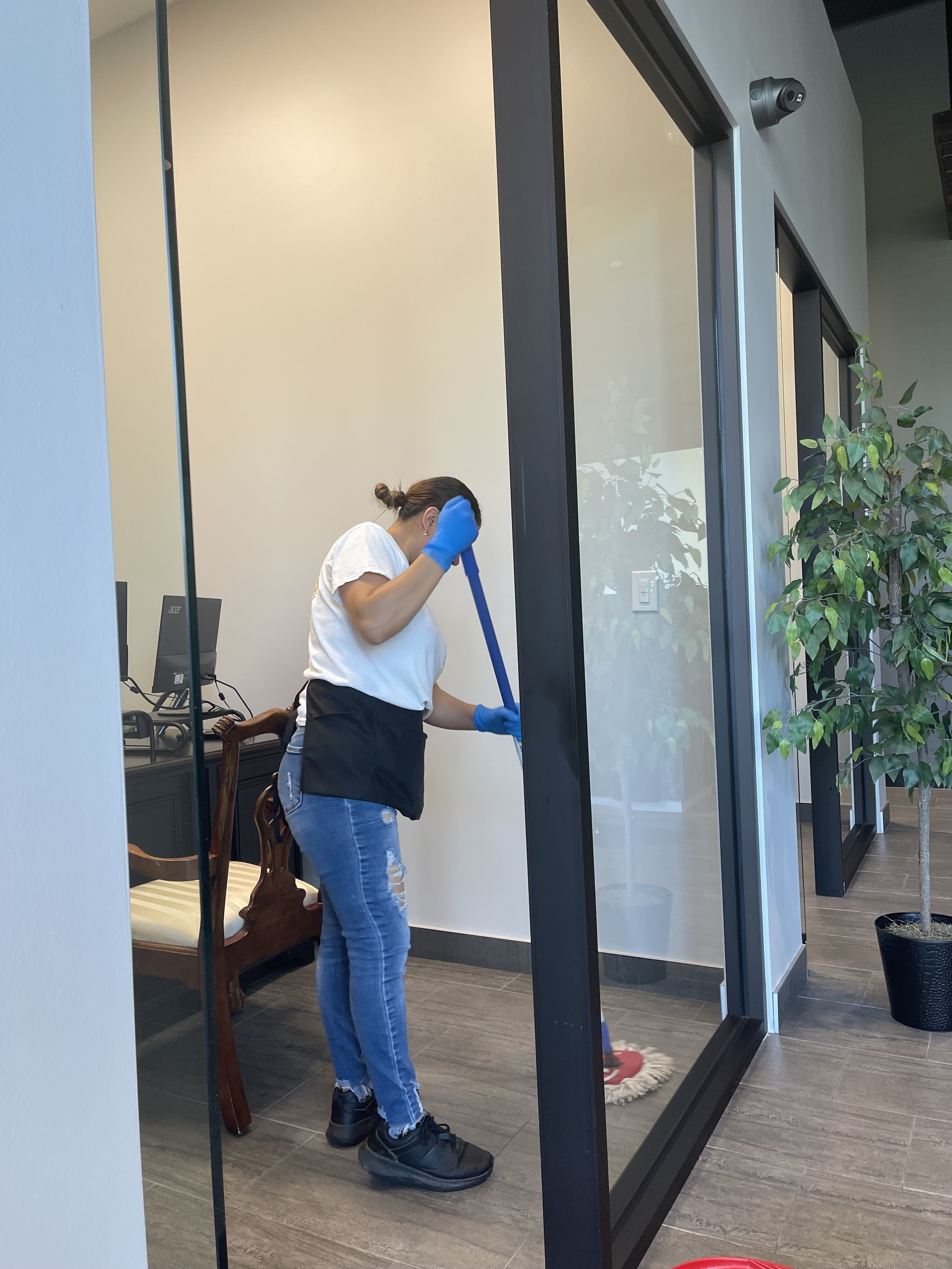 A person wearing blue gloves mops the floor in an office space with glass walls and a potted plant.