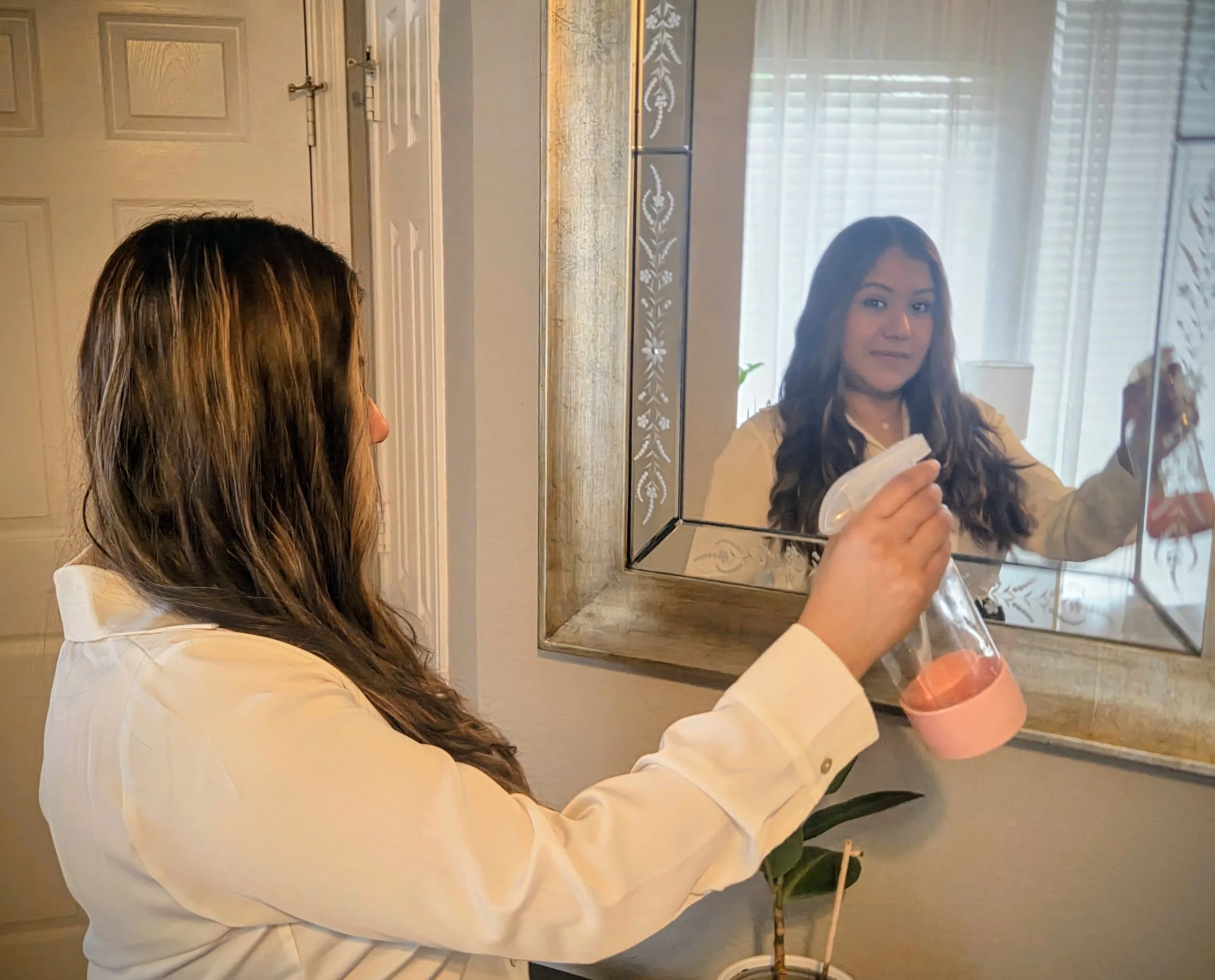 A woman with long brown hair wearing a cream-colored shirt, holding a spray bottle, is looking into a decorative mirror while standing in a well-lit room with a window in the background.