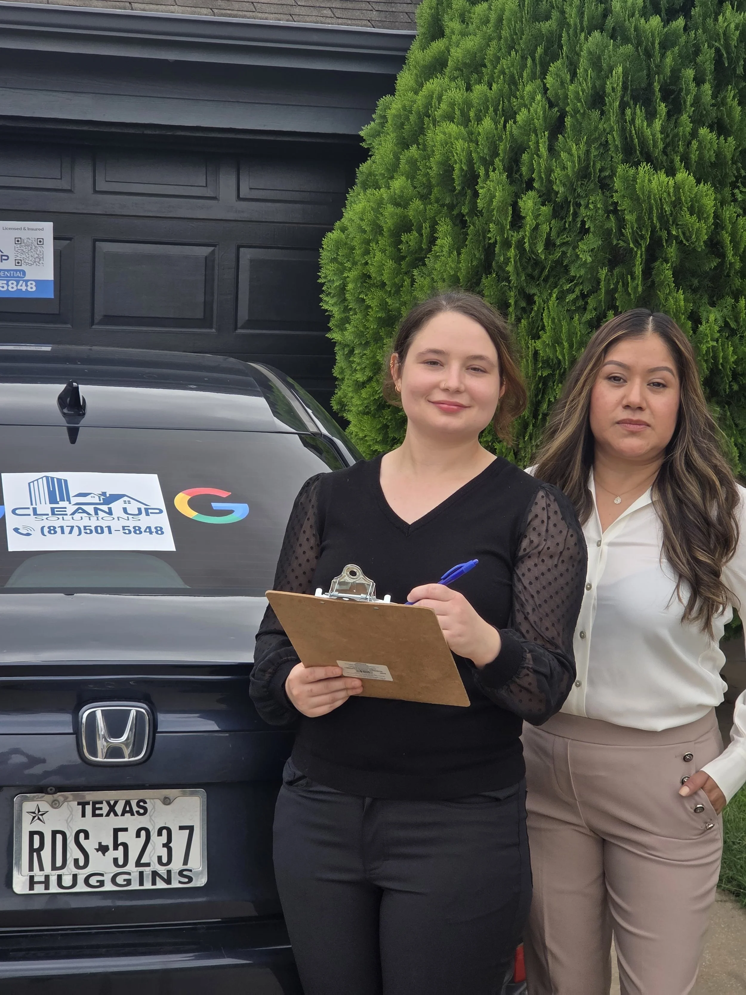 Two women standing in front of a black Honda vehicle with a sign on the back that says 'Clean Up' and a phone number. One woman is holding a clipboard and pen, smiling at the camera, while the other woman stands beside her, with a neutral expression.