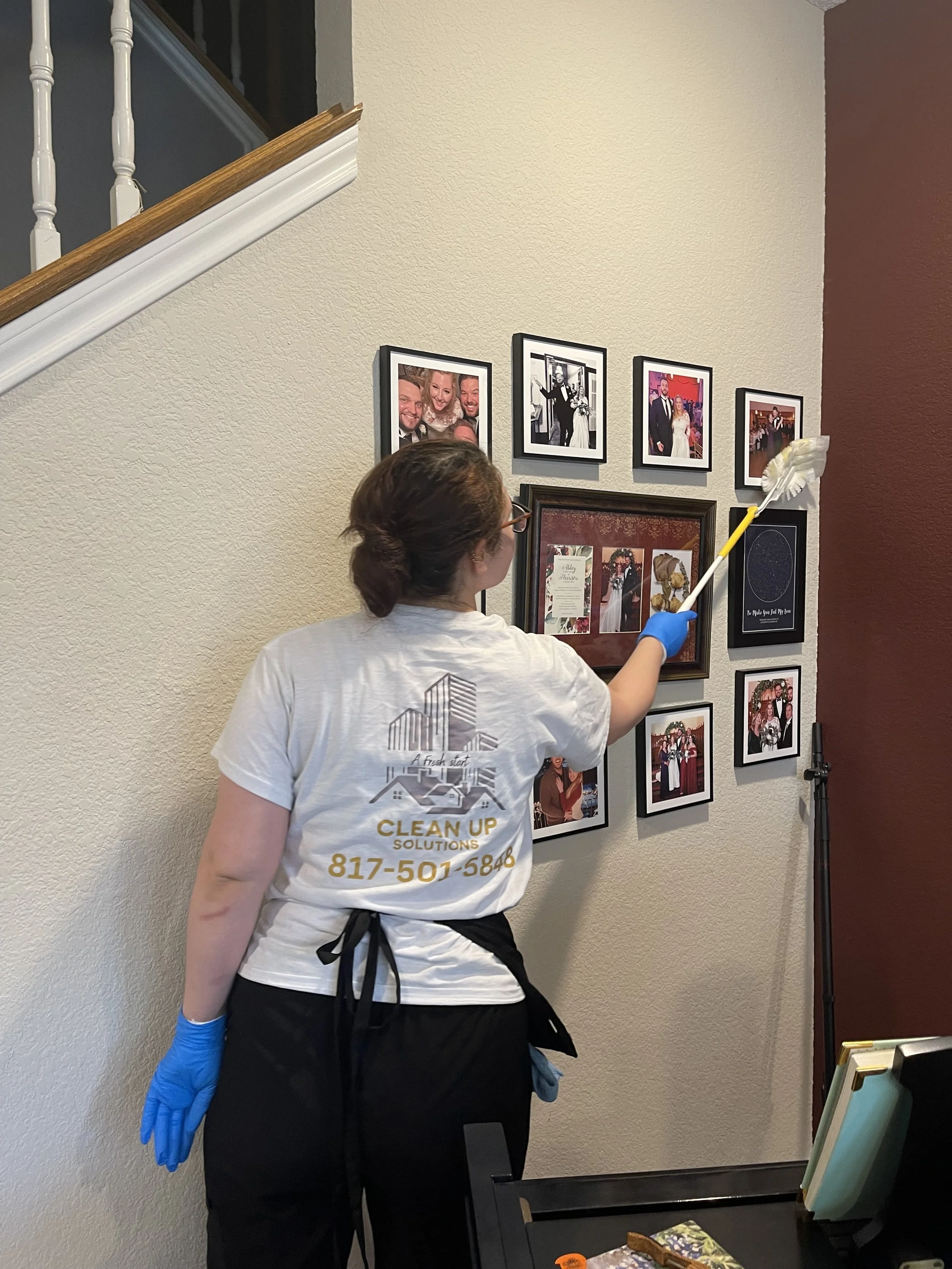 A woman cleaning a wall with framed photographs at a wedding or celebration, holding a duster with a long yellow pole, wearing blue gloves, black apron, light gray shirt, and glasses.