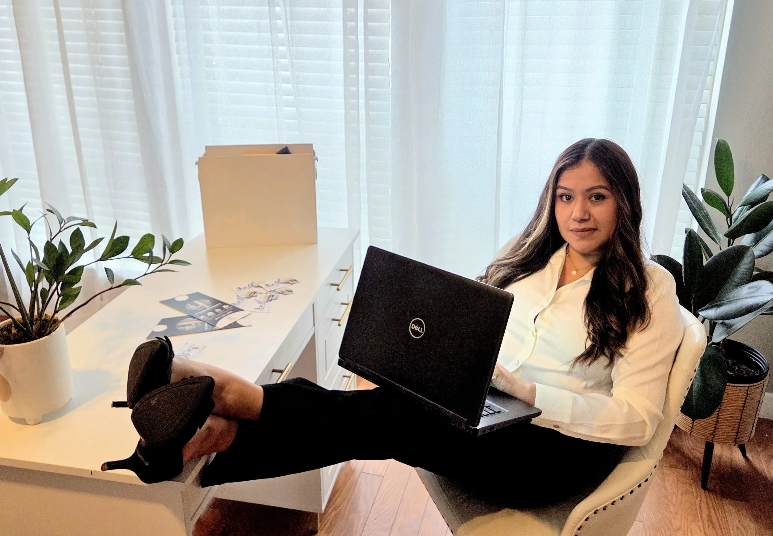Woman sitting at a desk with her feet up on the table, holding a laptop, in a well-lit room with plants and cash scattered on the desk.