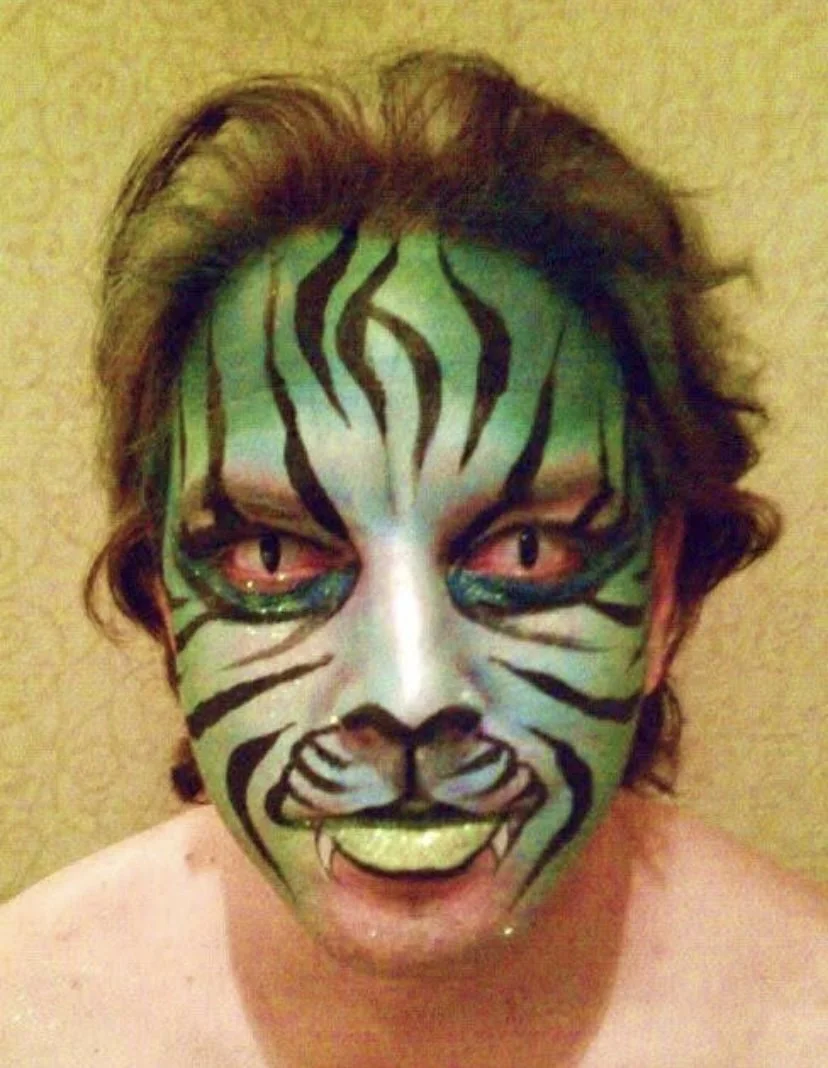 A person with green face paint and black tiger stripe patterns, having yellow eyes, red sclera, and fangs, resembling a fierce tiger.