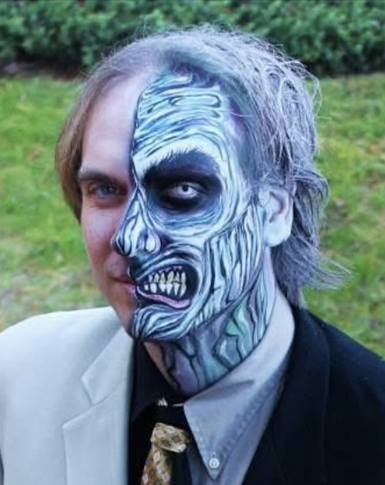 A man with half of his face painted as a blue zombie with exposed skull, sharp teeth, and hollow eye socket, standing outdoors on a grassy area.