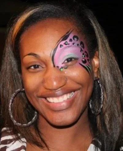 Woman with a pink and purple butterfly face paint design over her left eye, smiling, wearing hoop earrings.