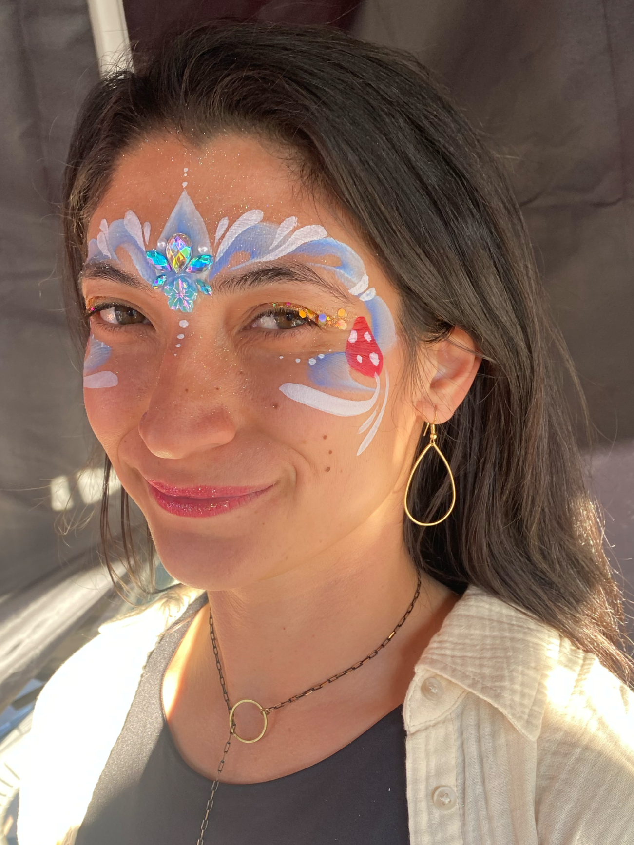 A woman with face paint featuring blue, white, and red colors, including a decorative jewel on her forehead, and glitter, smiling with earrings and necklaces.