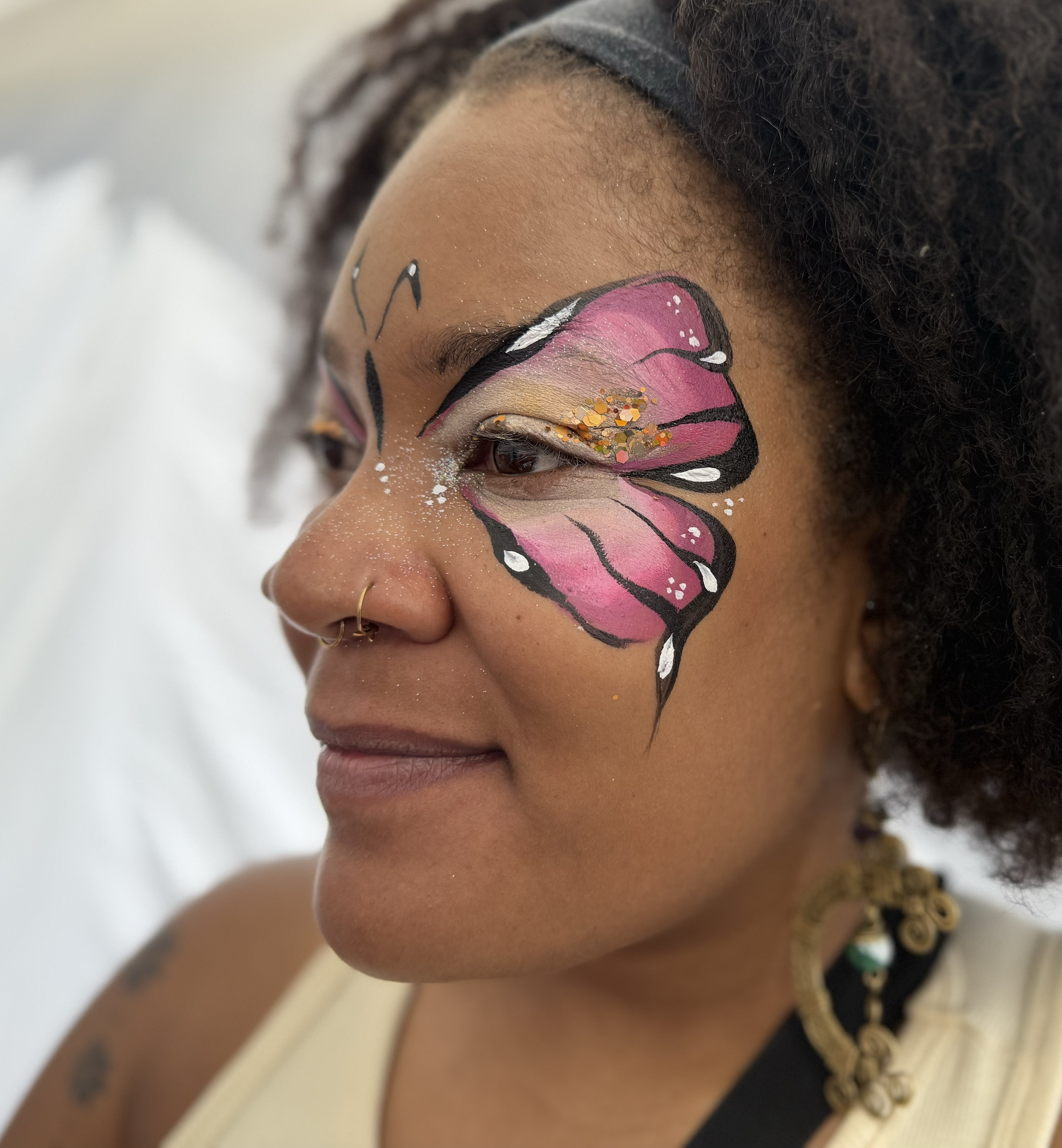 A woman with a butterfly face painting on her right cheek, wearing gold jewelry and a black headband, with a blurred background.