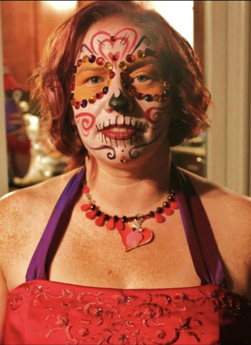 Woman with sugar skull face paint decorated with red and black rhinestones, red and pink lips, and a large heart-shaped pendant necklace, wearing a red embroidered dress with purple straps, indoor setting with warm lighting.