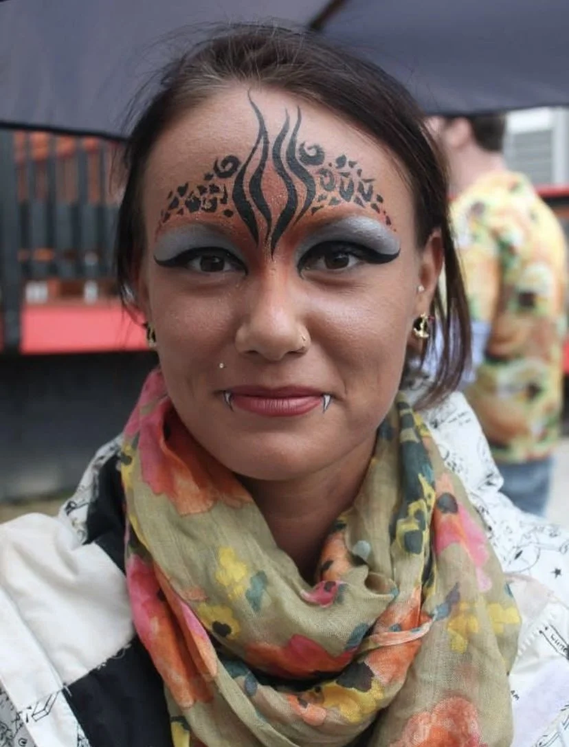 Woman with elaborate black face paint design and bold eye makeup, wearing earrings, a nose piercing, a lip piercing, and a patterned scarf.