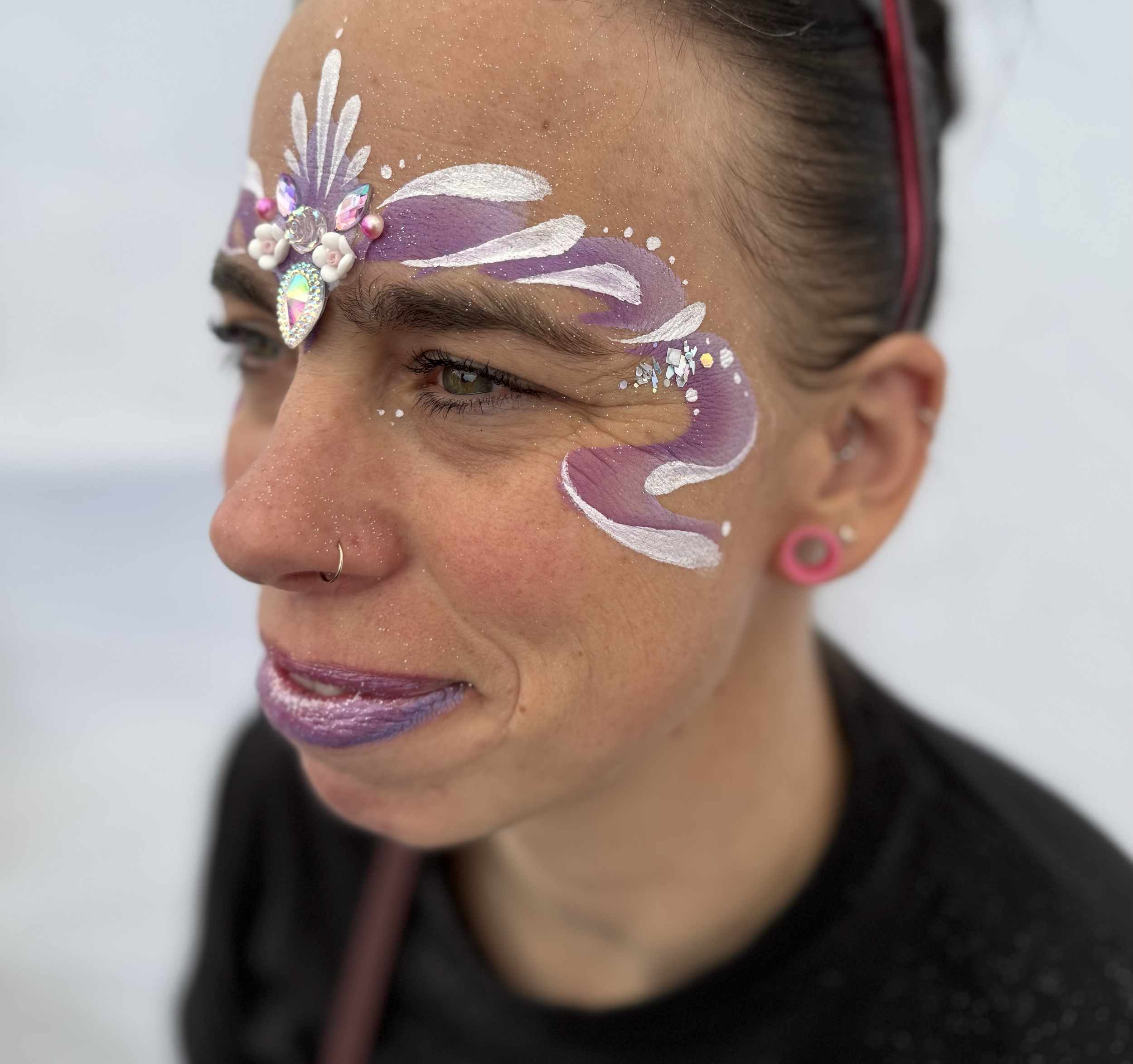 A woman with elaborate face paint, including purple, white, and silver designs, rhinestones, and decorative gems above her eyebrow, wearing a pink earring and a nose ring, with her hair pulled back.