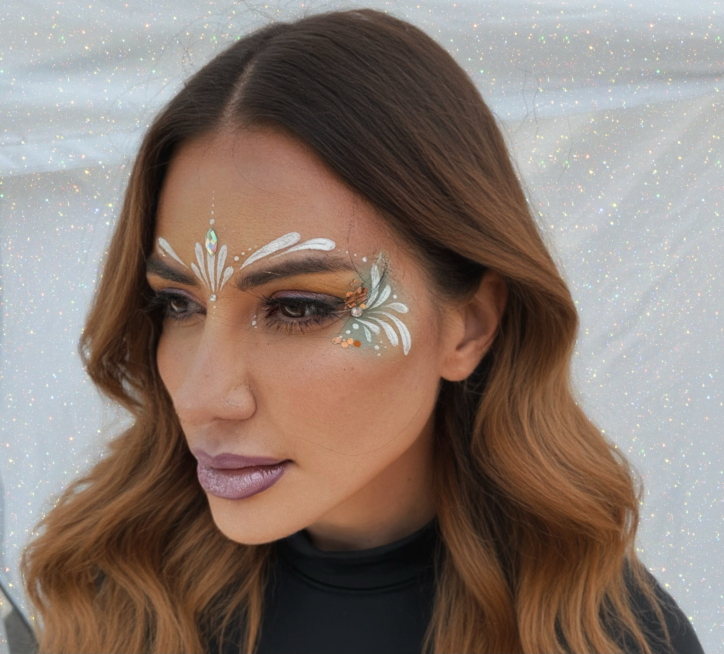 A woman with long, wavy brown hair, wearing makeup and silver lipstick, has intricate face paint with white, blue, and orange accents resembling butterflies on her face, with a glittery background.