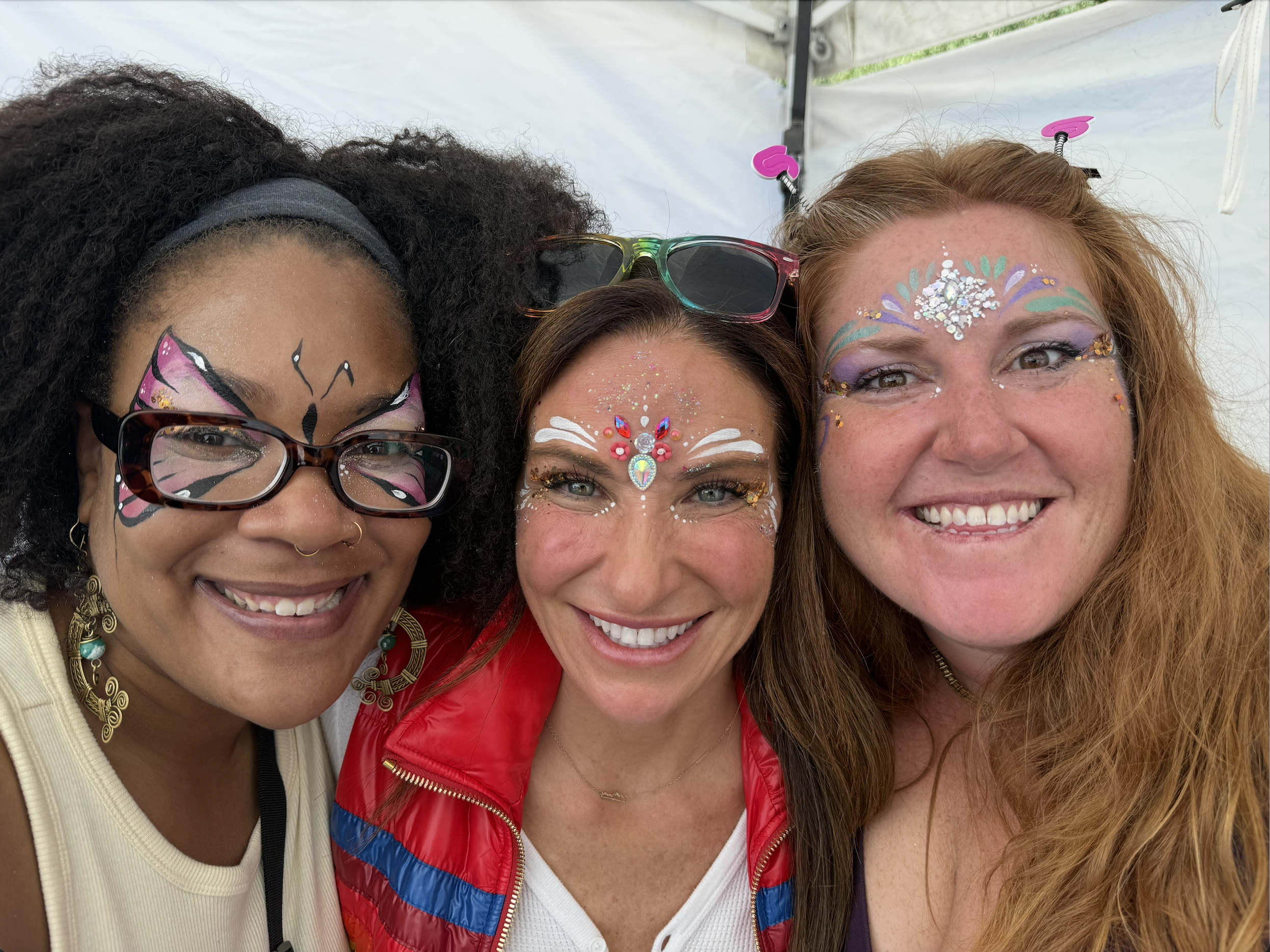 Three women with colorful face paint and glitter smiling together in a close-up selfie, outdoors under a tent.