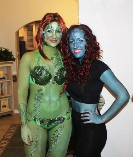 Two women in costumes with body paint; one with green body paint and red hair, the other with blue body paint and red curly hair, posing indoors.