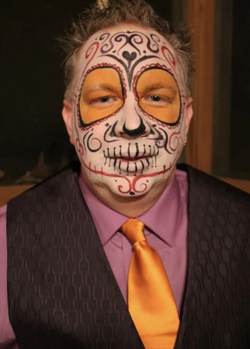 Person with sugar skull face paint featuring orange eye blocks, black and red decorative details, and wearing a purple shirt with an orange tie.