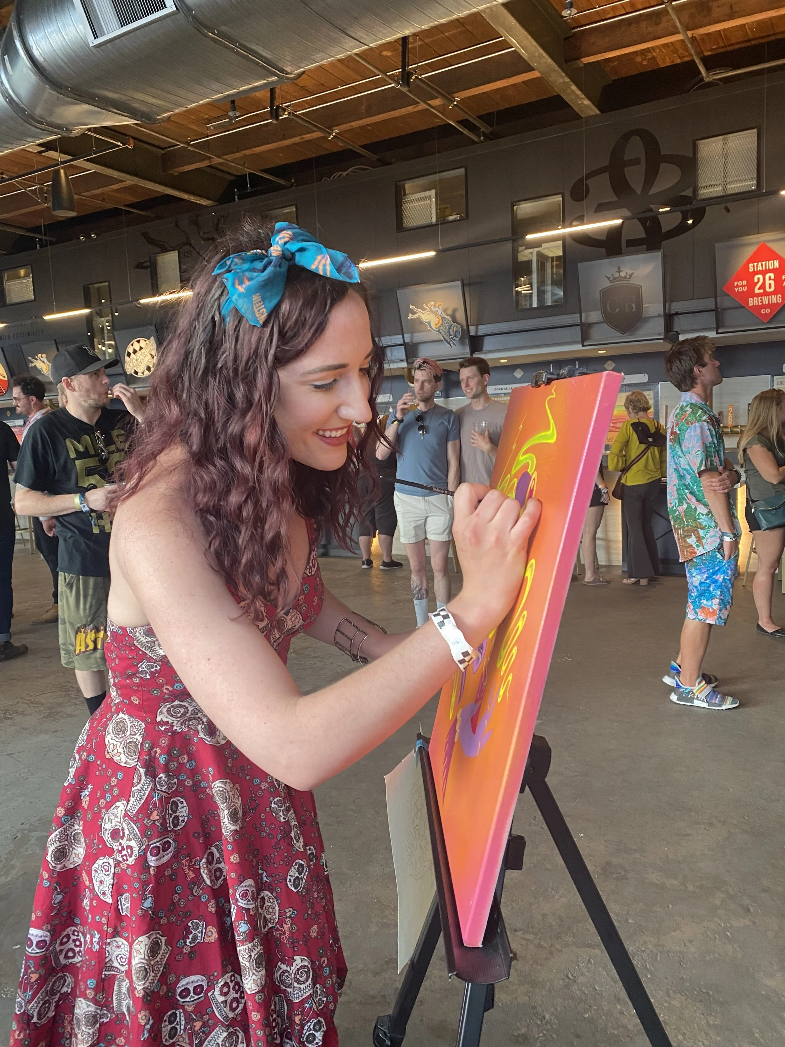Young woman with curly hair wearing a blue headband and a red dress with skull prints, smiling as she paints on a colorful canvas at an indoor event. People in the background are mingling and taking pictures.