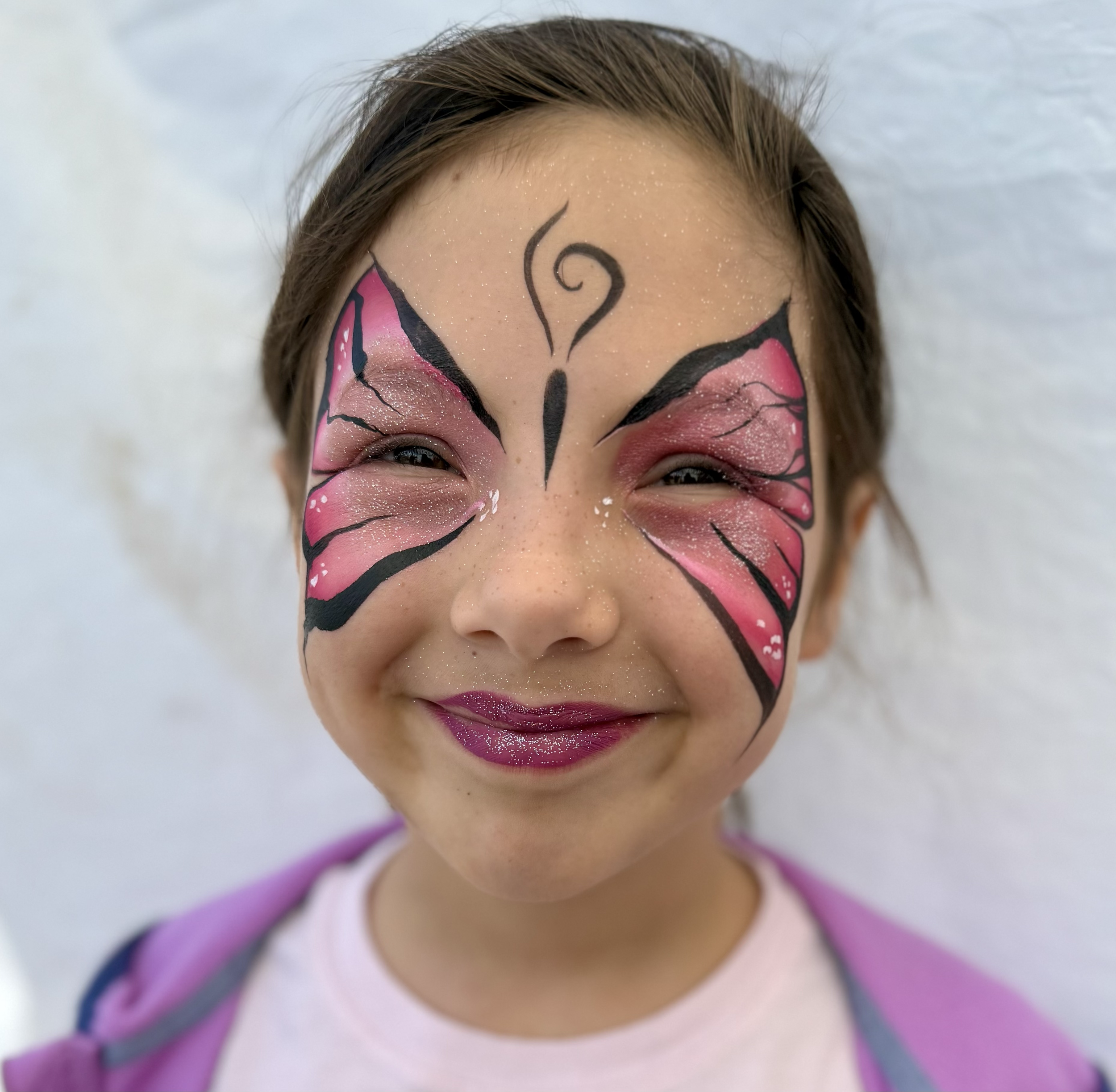 Child with pink and black butterfly face paint, glitter on face, smiling with lips painted purple, wearing a pink shirt and purple jacket, standing against a white wall.