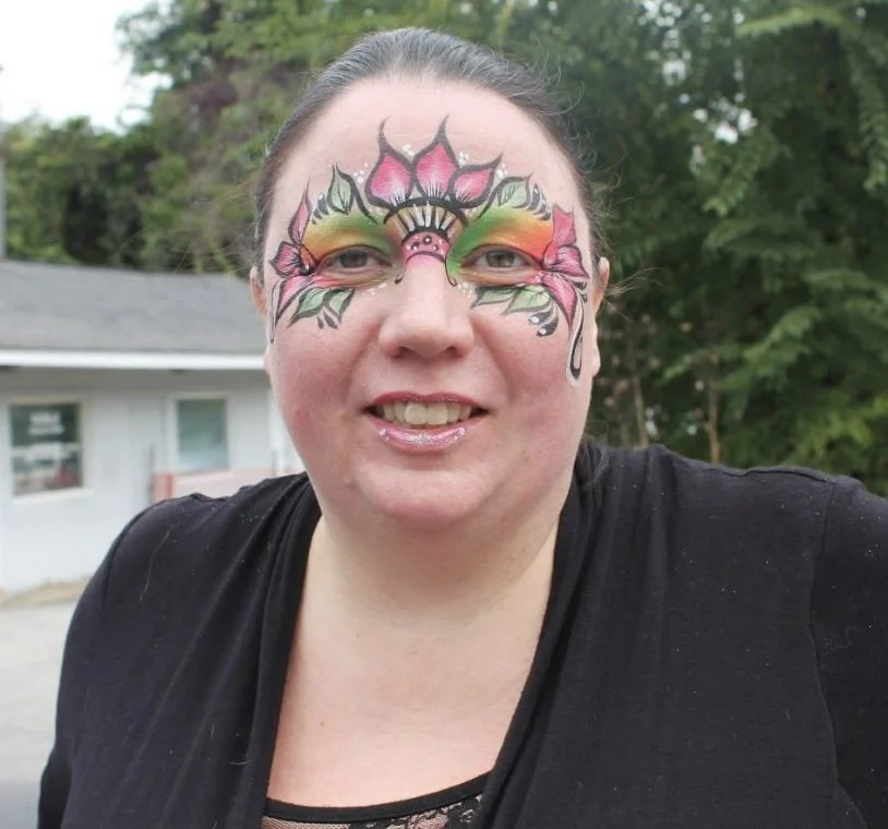 Woman with colorful floral and leaf face paint around her eyes, smiling outdoors with trees and a house in the background.