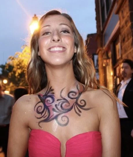 Young woman with long wavy hair smiling outdoors at sunset, wearing a pink strapless dress and having a large floral tattoo on her chest.