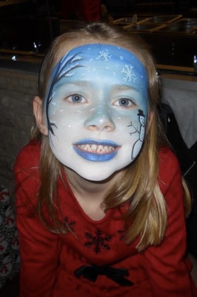A girl with face paint depicting a winter scene, including snowflakes and a small penguin, wearing a red shirt with black details.