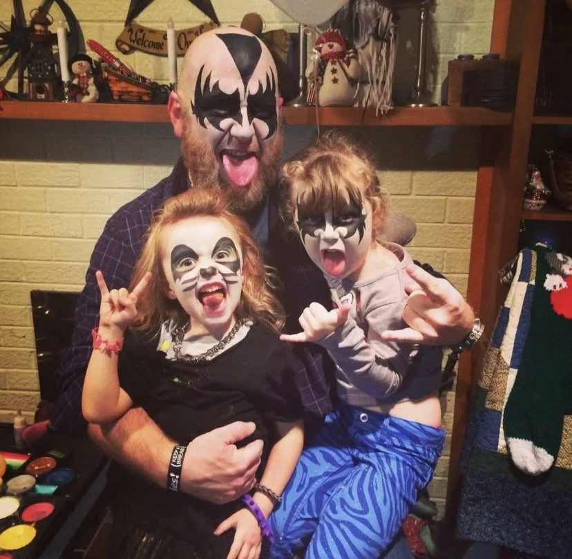 A man and two young girls with face paint, making rock hand gestures, all smiling and having fun at a festive indoor gathering.