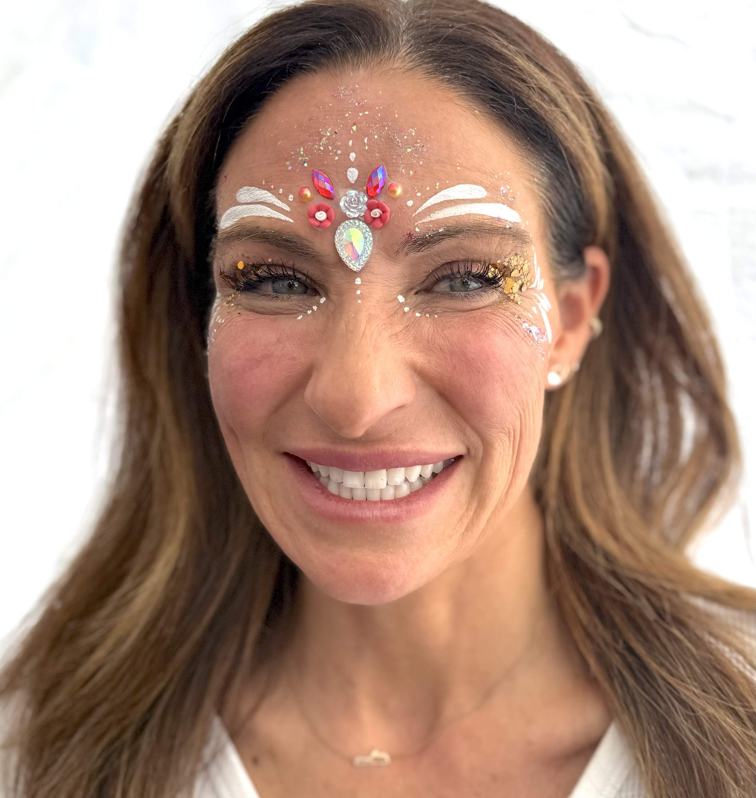 A woman smiling with face decorated in colorful rhinestones, paint, and glitter, including a large jewel on her forehead and white decorative patterns around her eyes and forehead.