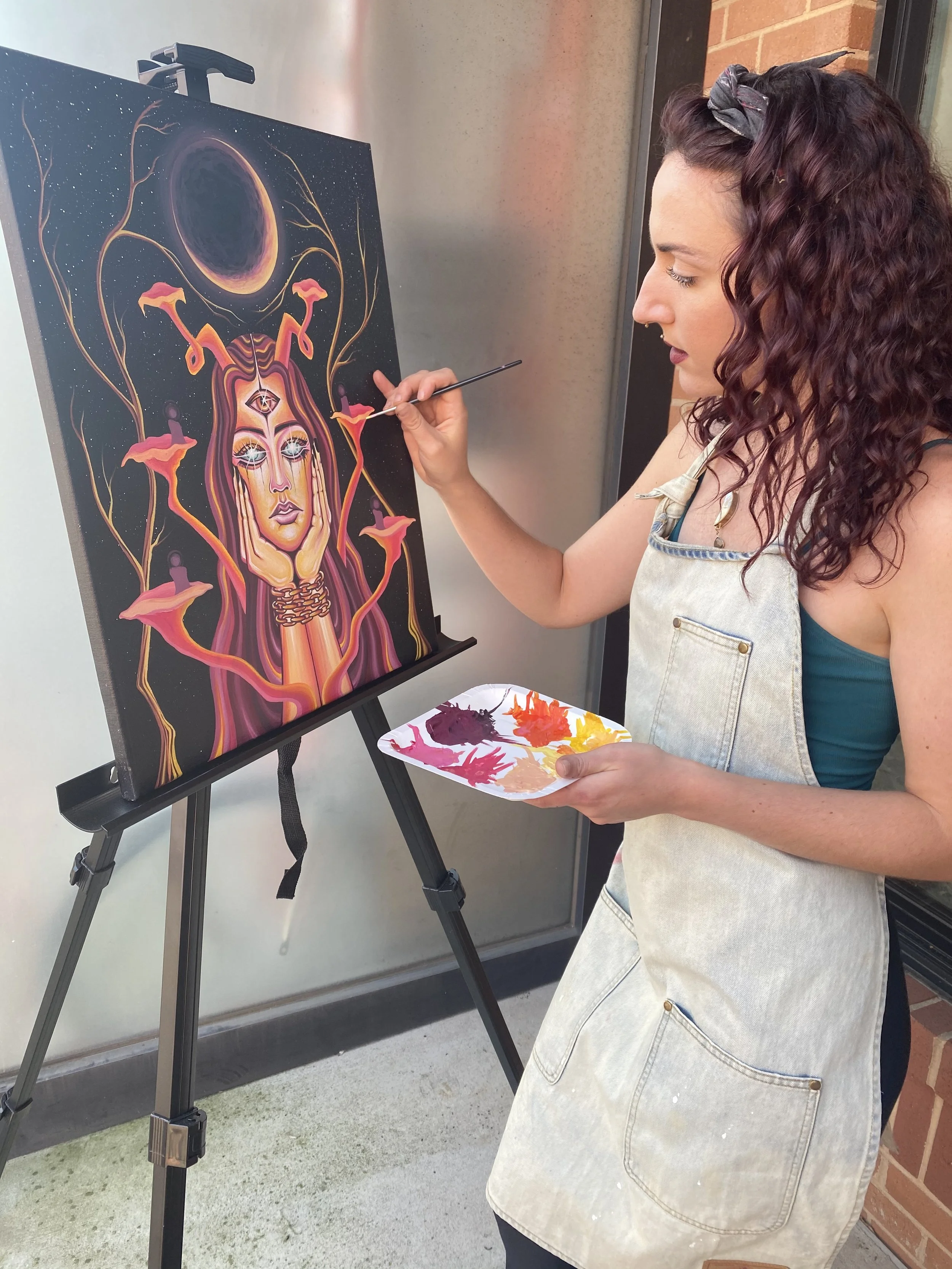A woman painting a surreal portrait of a woman with long hair, multiple eyes, and candles on her head, on a black canvas with a cosmic background, standing outside near a brick wall.