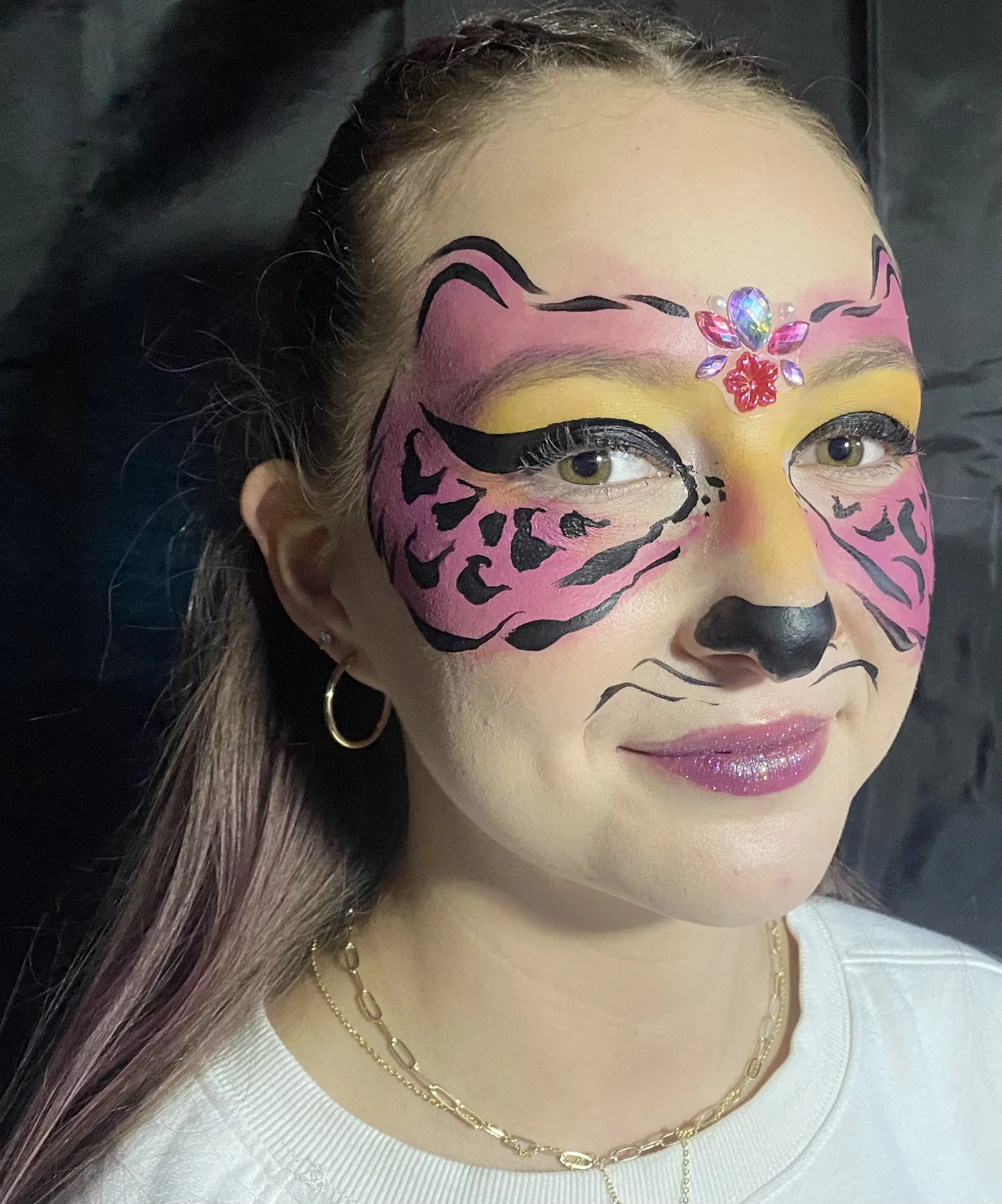 A woman with tiger face paint, yellow eyeshadow, glittery pink lipstick, wearing a white top, gold chain necklace, earrings, and with decorative rhinestones on her forehead.