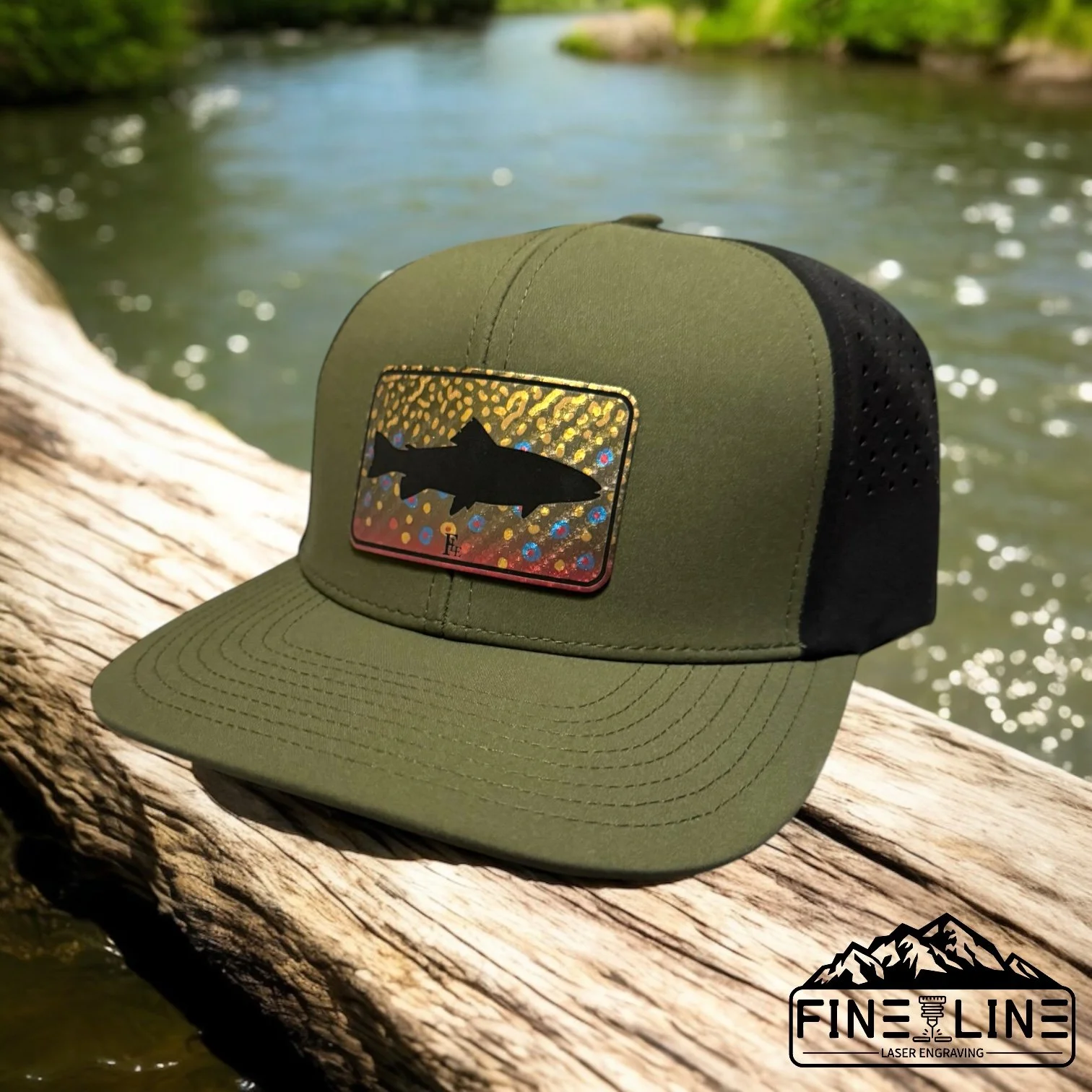 Green and black trucker hat with a colorful fish logo and laser-engraved mountain logo, resting on a weathered piece of wood near a body of water