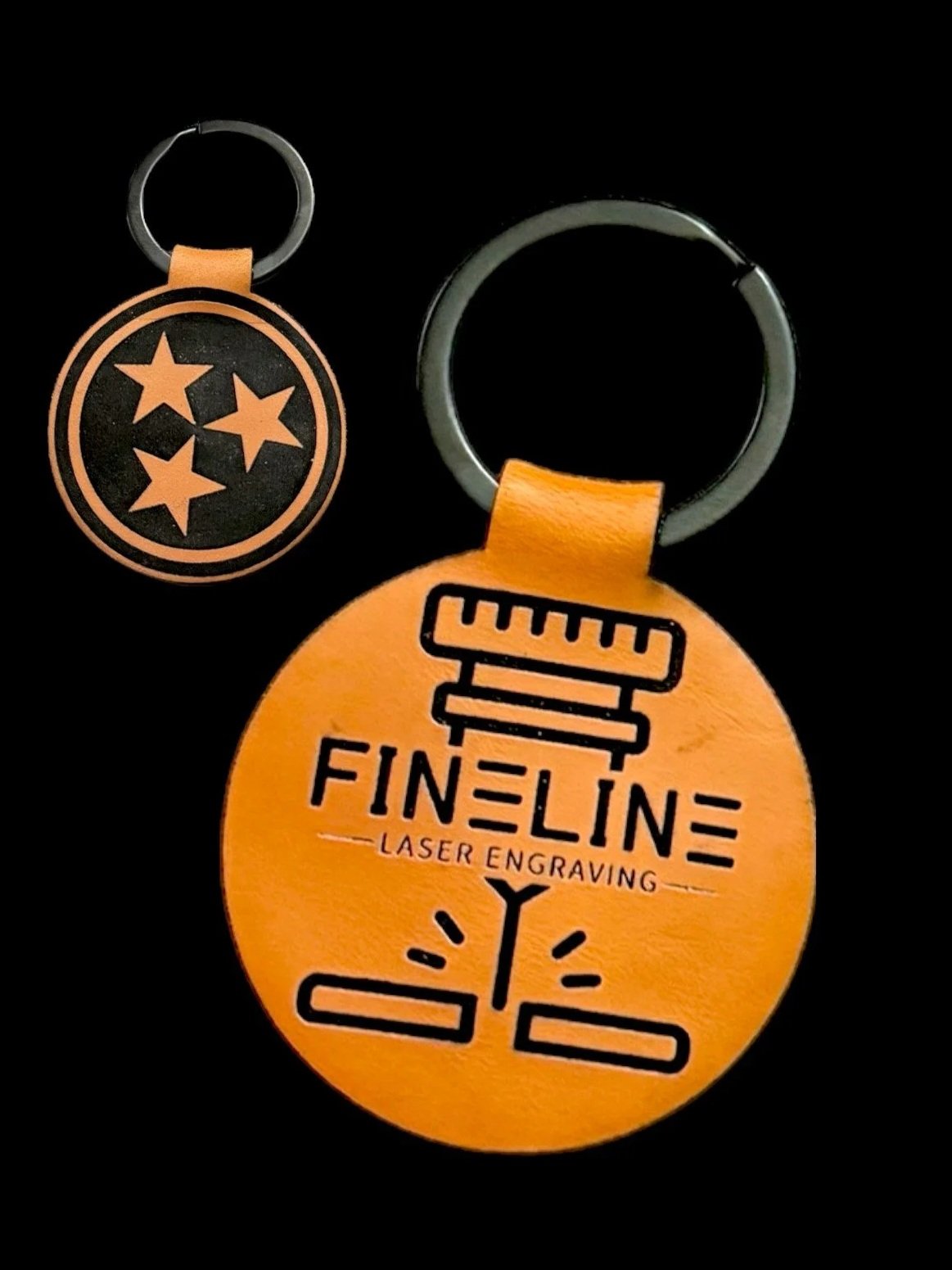 Two orange circular keychains with black designs, one featuring the Tennessee state symbol and the other with text 'FINE LINE Laser Engraving' and graphic elements, on a black background.