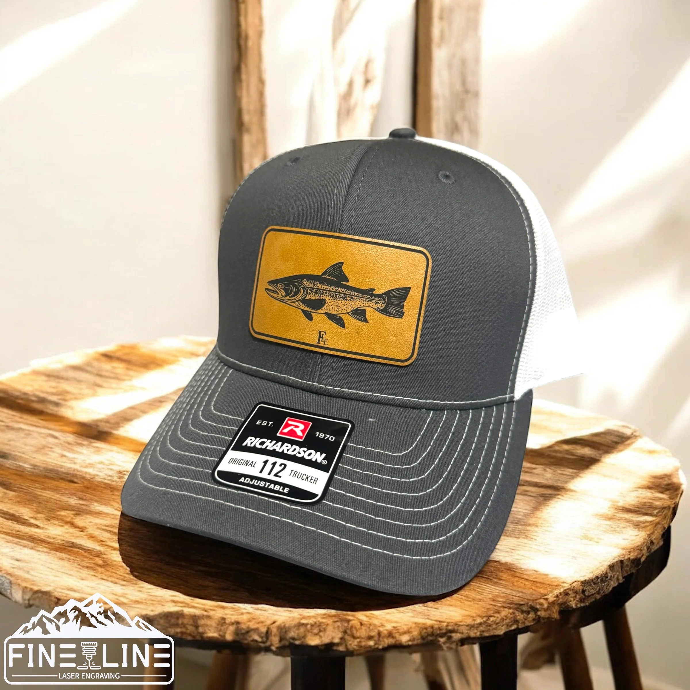 A gray and white Richardson trucker cap with a yellow patch featuring a fish illustration, placed on a wooden surface.