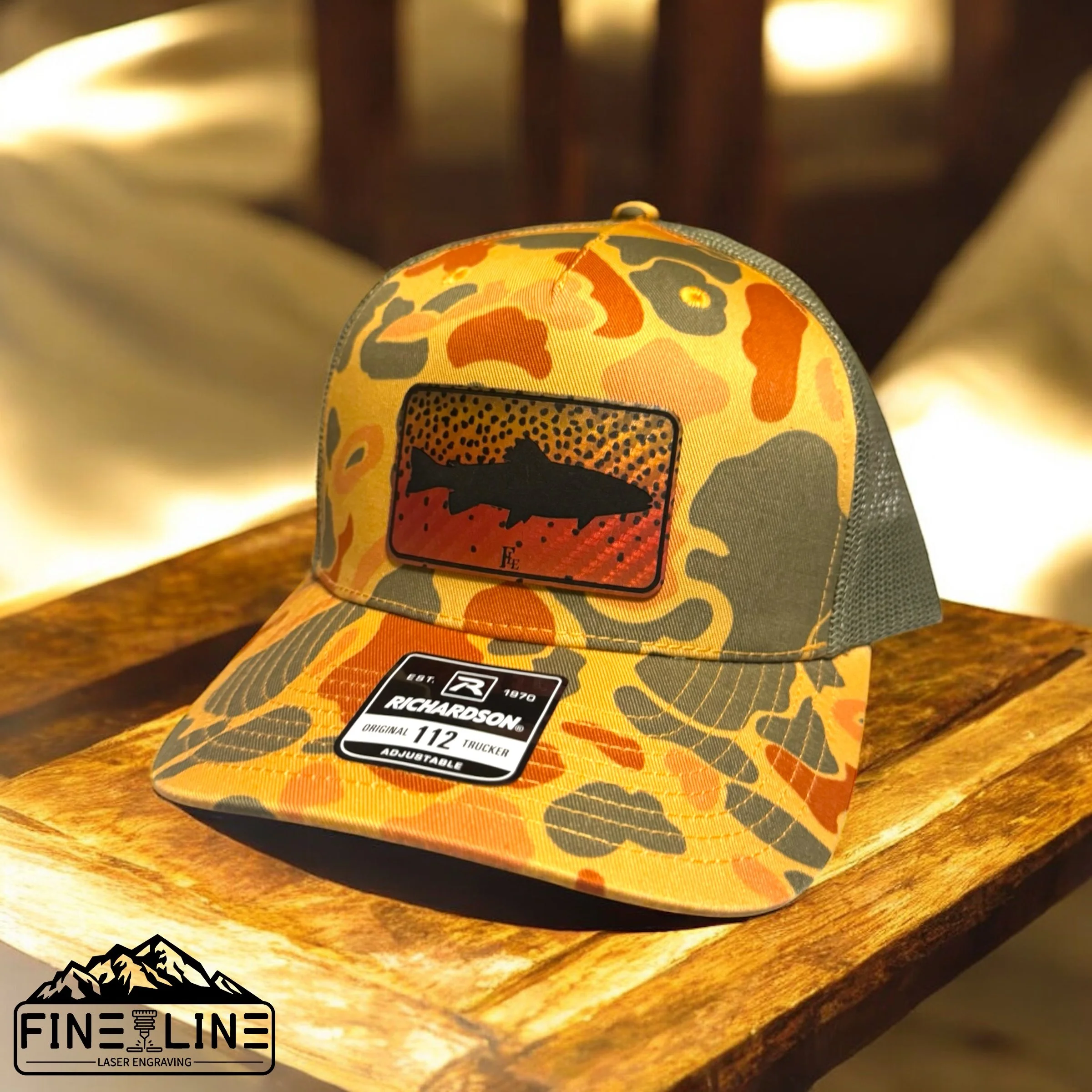 Camouflage cap with a black fish silhouette patch and Richardson brand sticker resting on a wooden surface.