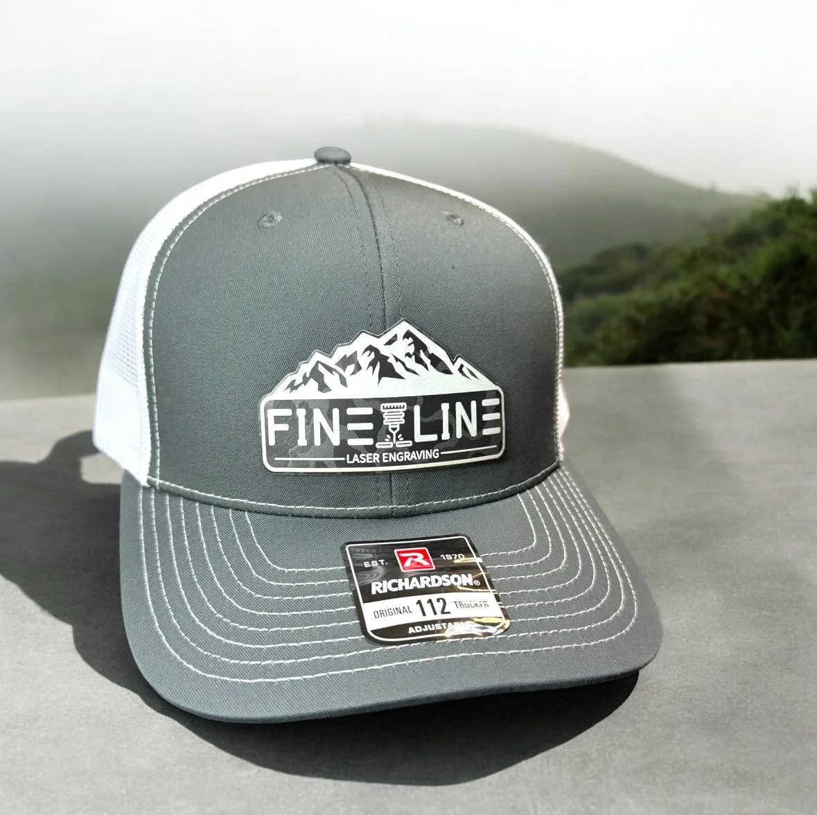 Gray trucker hat with Mountain logo and 'FINE LINE LASER ENGRAVING' text on front, placed on a flat surface outdoors with foggy mountains in the background.