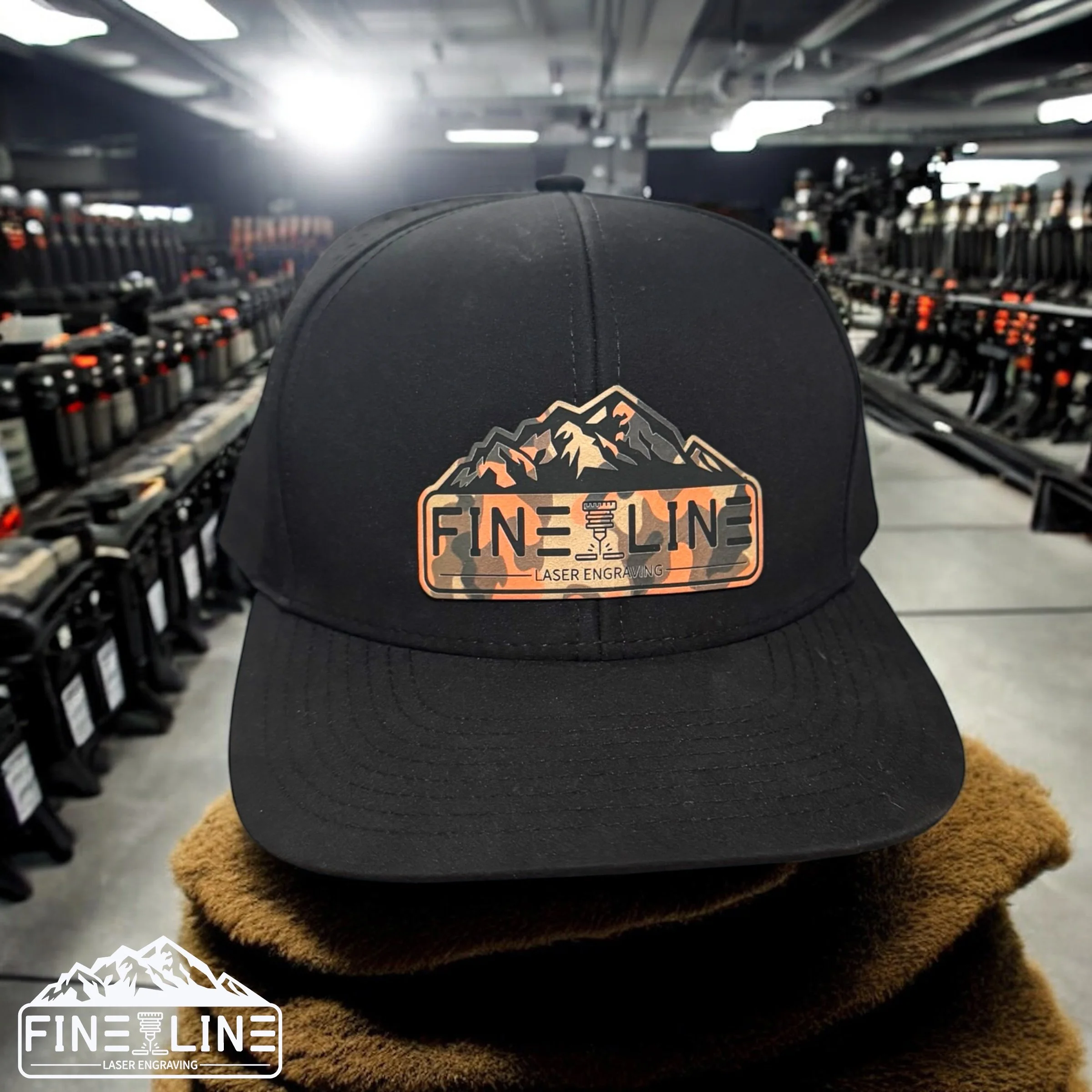 A black baseball cap with a mountain graphic and the text "FINE LINE" and "LASER ENGRAVING" on the front, displayed on a brown surface in a retail store with rows of hardware items in the background.