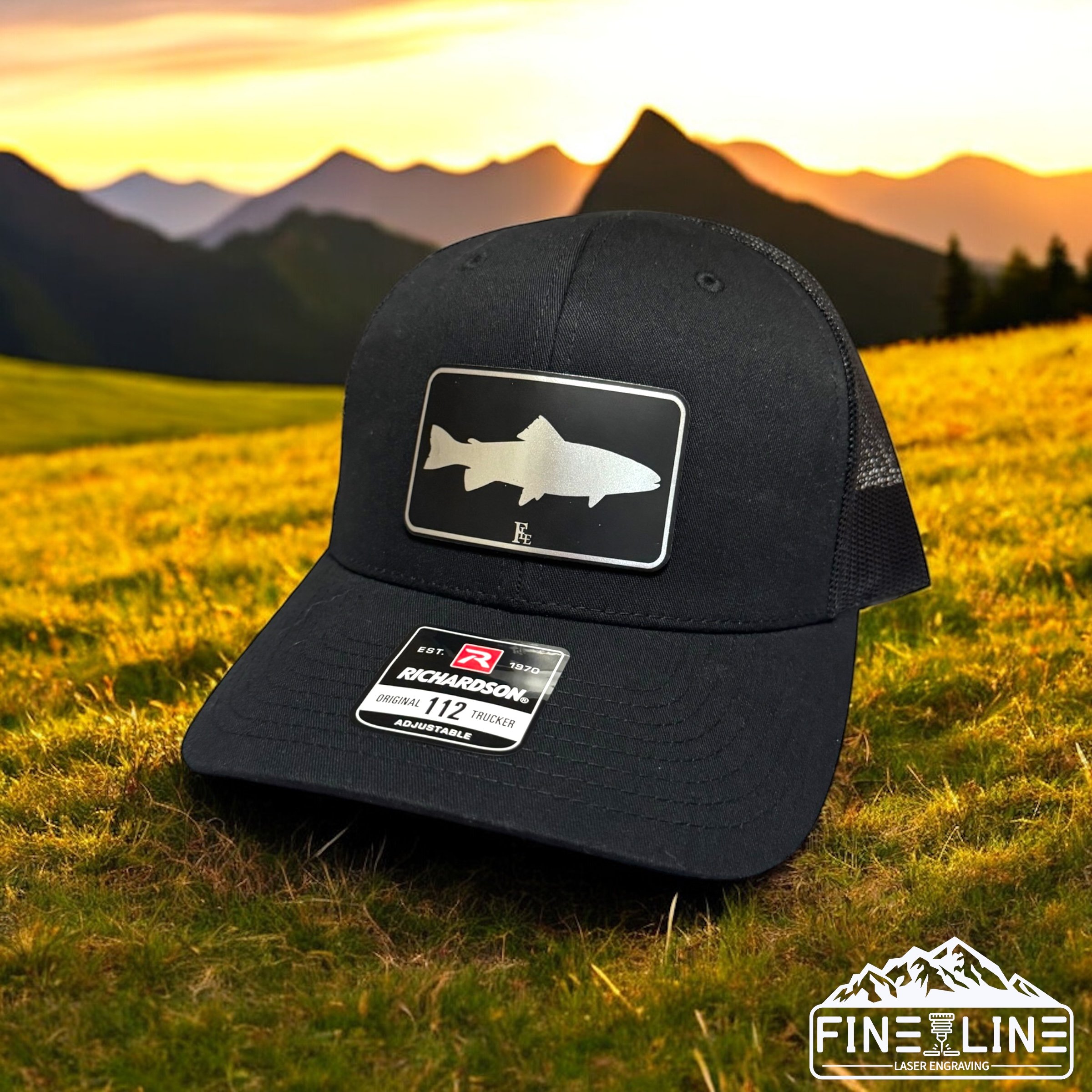 Black trucker hat with a fish silhouette patch, placed outdoors on grass with mountains and sunset in the background.
