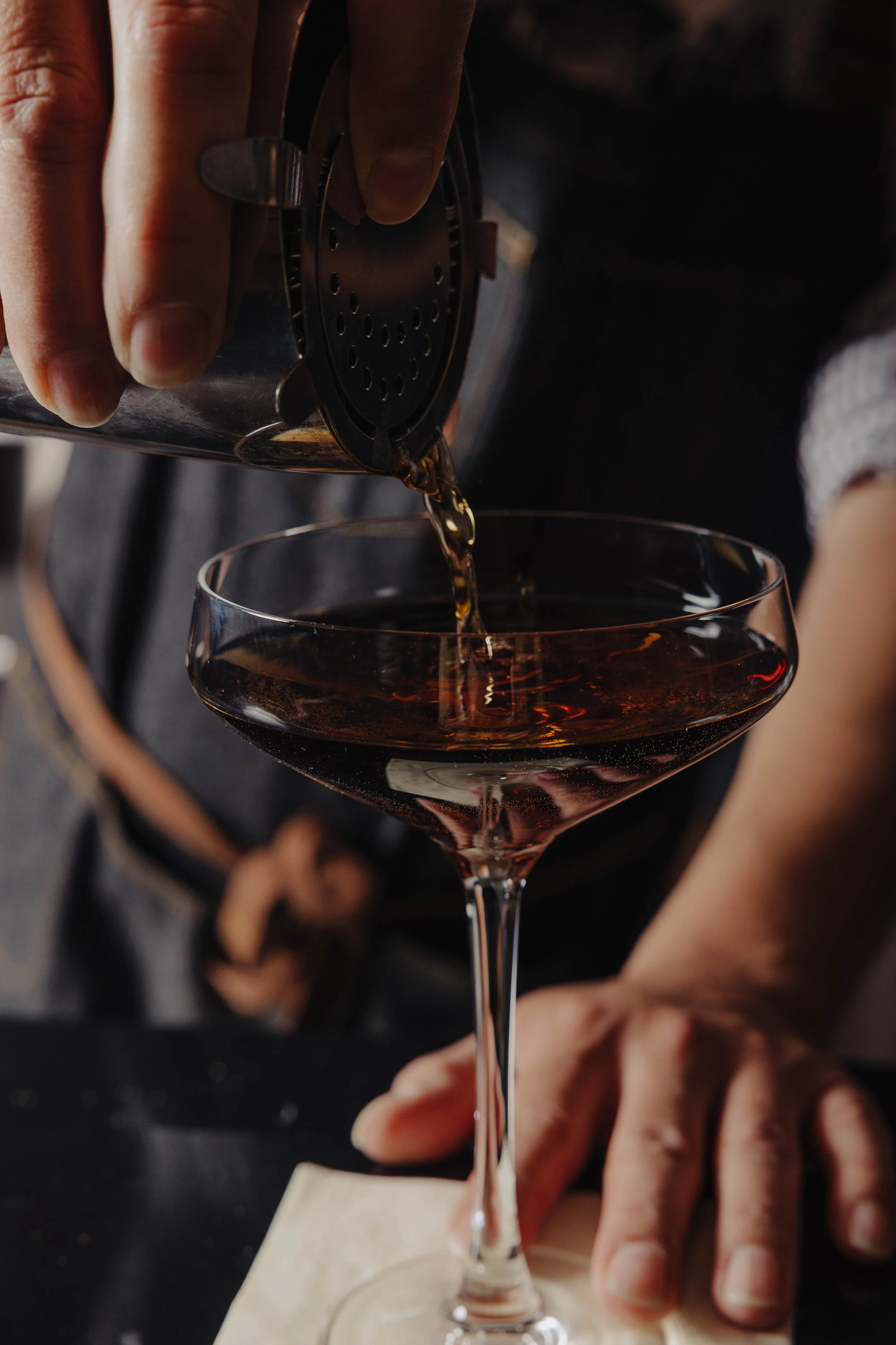 Close-up of a person pouring dark liquid, likely alcohol, from a shaker into a cocktail glass.
