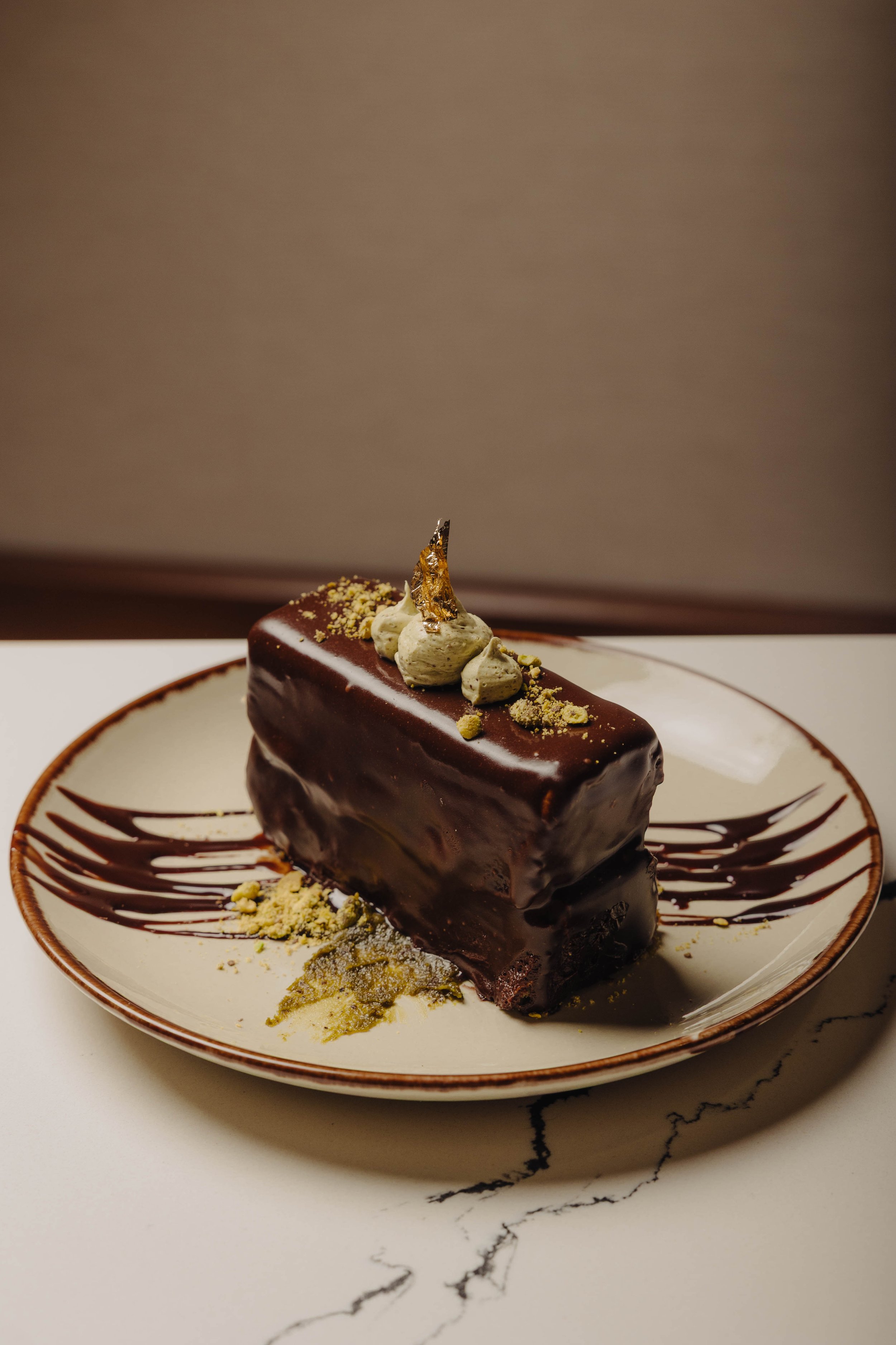 Chocolate mousse cake topped with whipped cream, gold leaf, and crushed pistachios, served on a plate with chocolate drizzle and gold dust.
