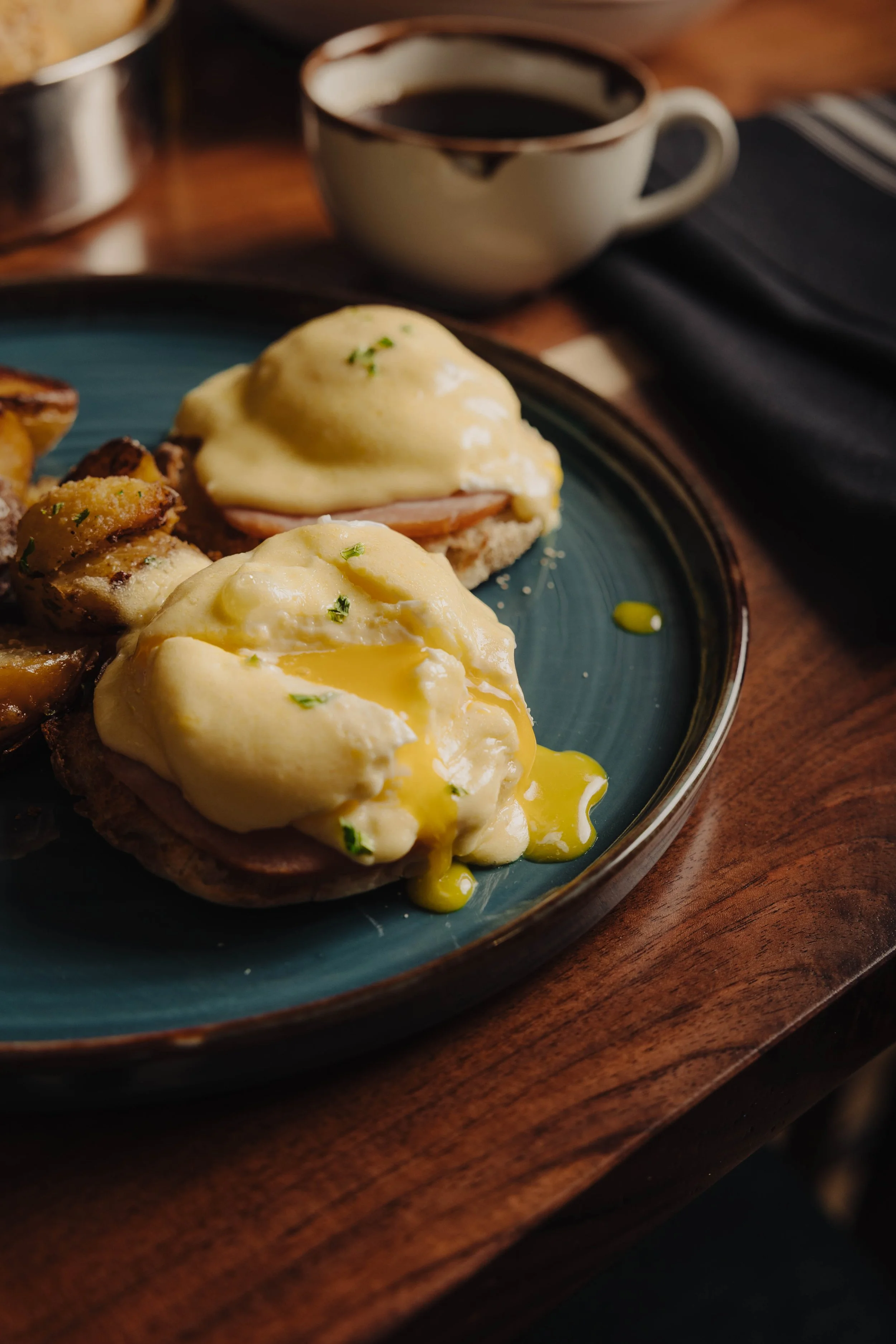 Close-up of eggs Benedict with poached eggs, ham, and hollandaise sauce on a blue plate, with roasted potatoes and coffee in the background.