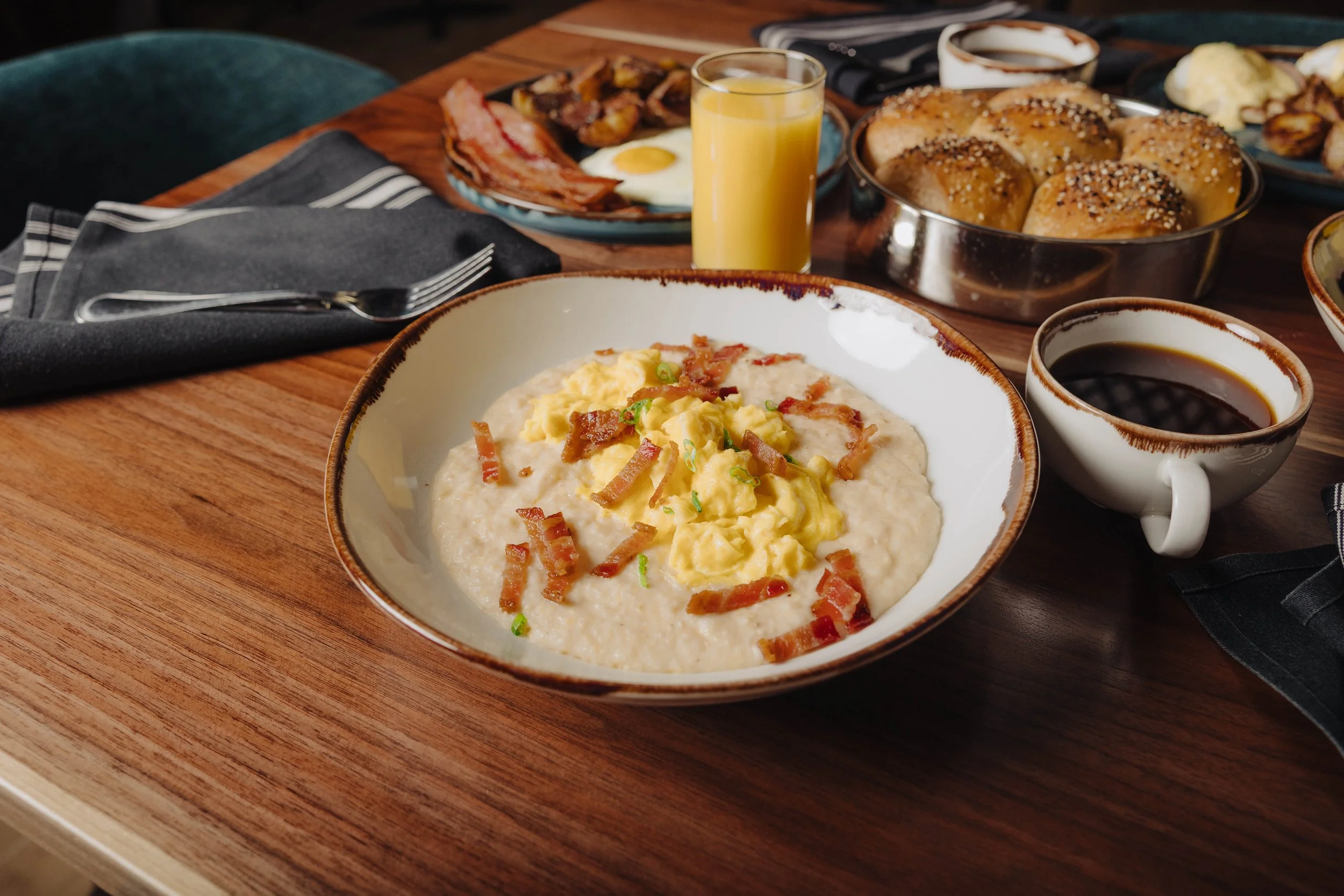 Breakfast with scrambled eggs topped with bacon, a cup of black coffee, a glass of orange juice, and various baked goods on a wooden table.