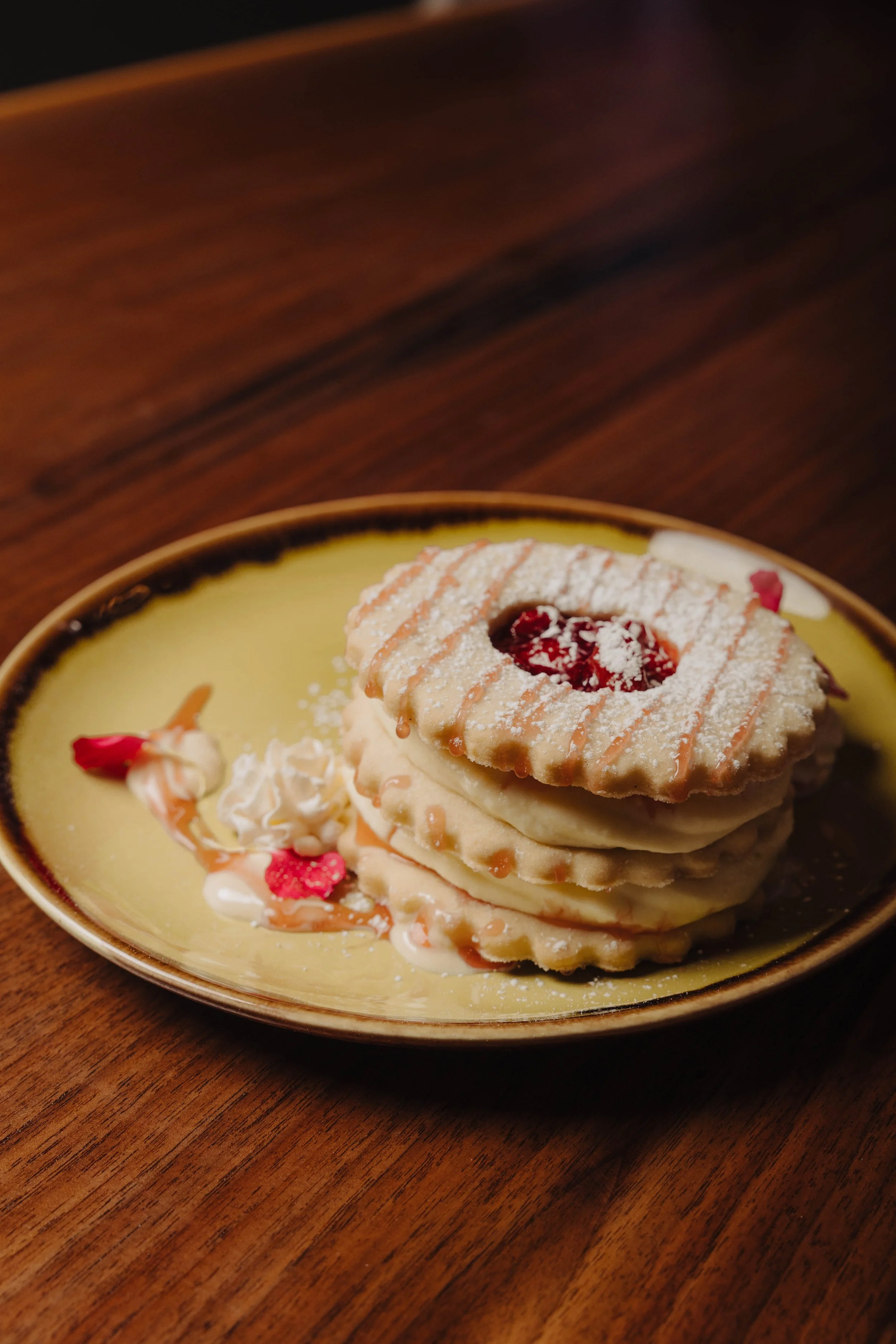Stack of three shortbread cookies with a jam center, served on a yellow plate with whipped cream, red flower petals, and caramel drizzle, on a wooden table.