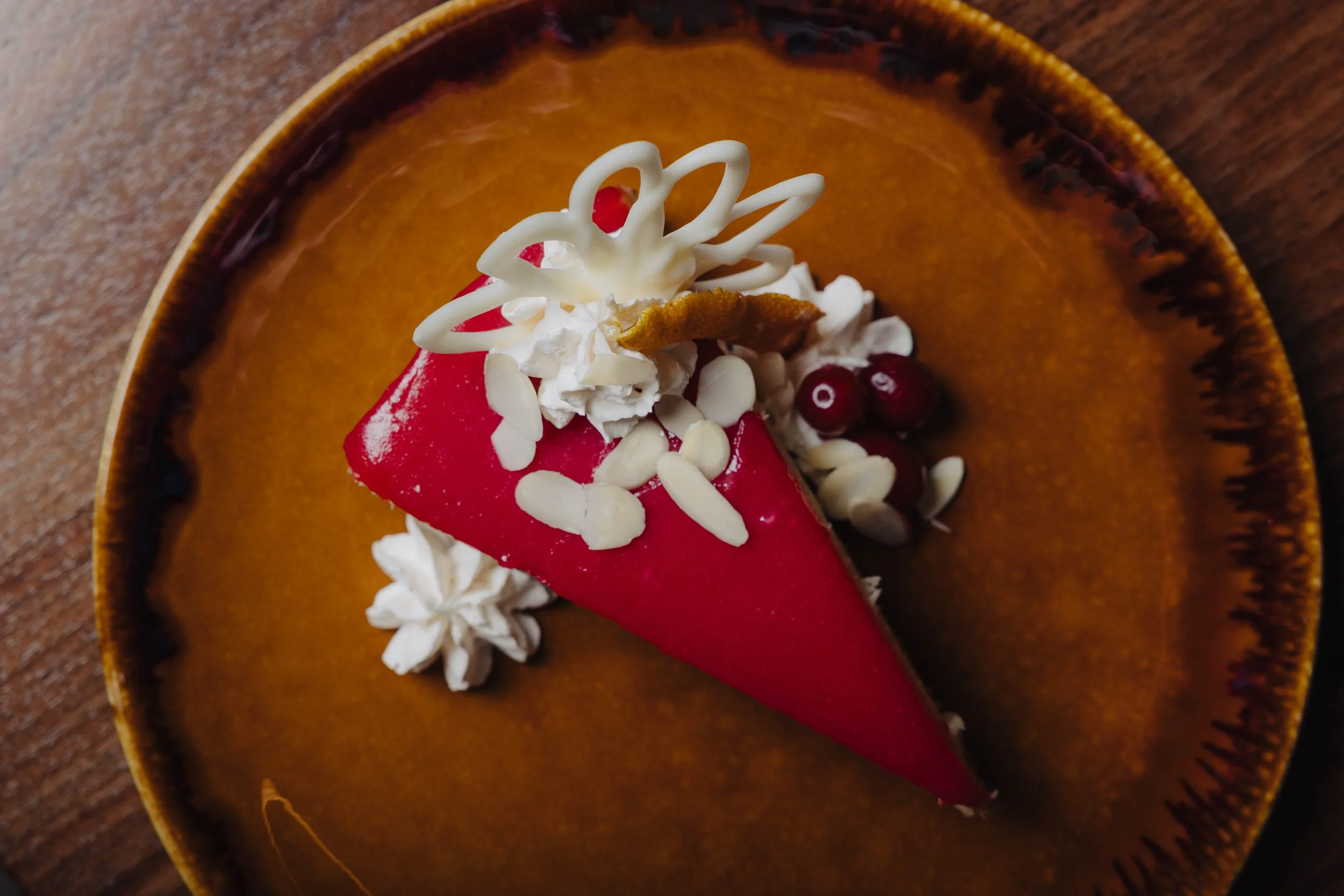A slice of red fruit cake with white icing, garnished with whipped cream, almond slices, red berries, and decorative white chocolate curls, served on a brown ceramic plate.