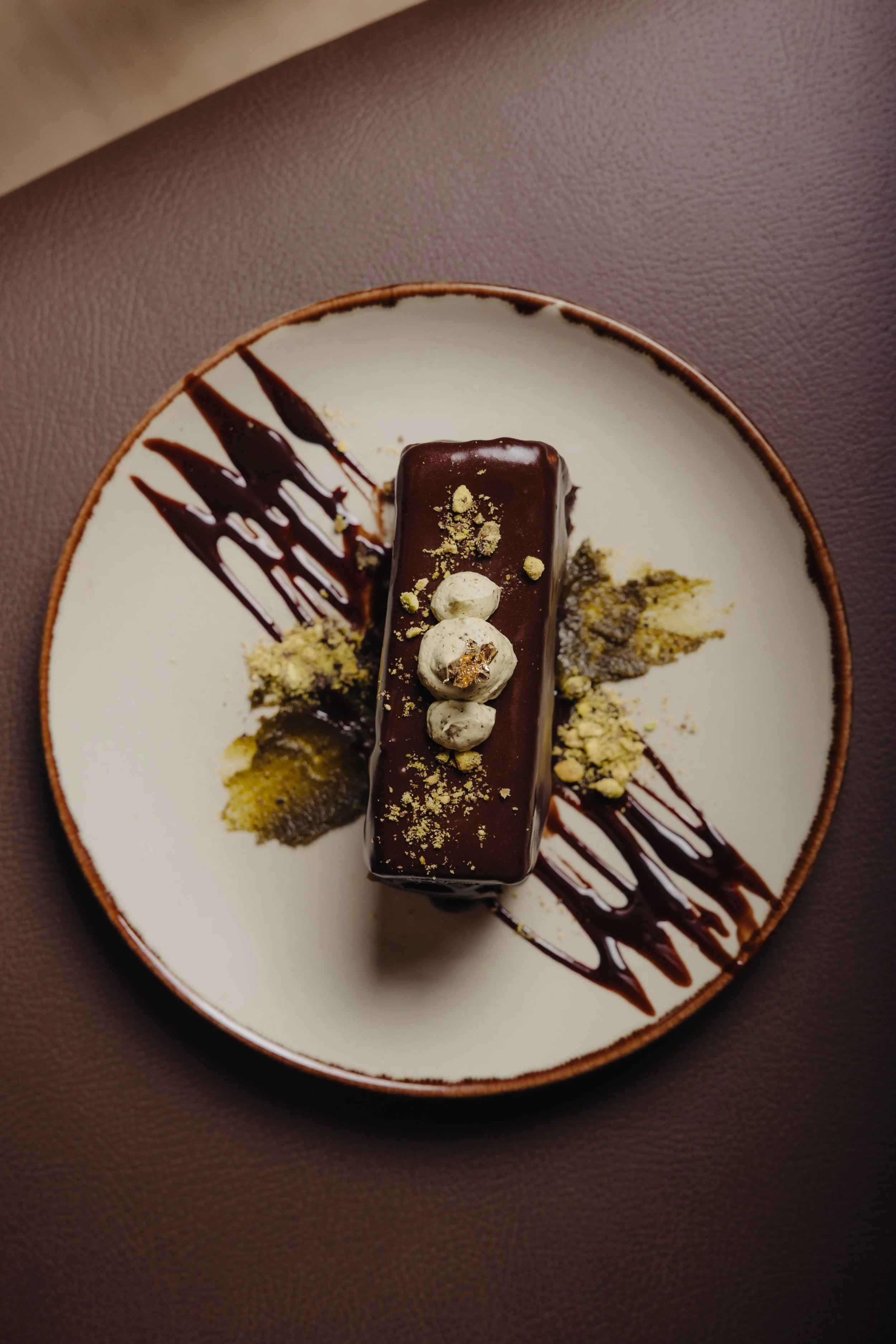 Chocolate dessert on a white plate with chocolate sauce, crushed nuts, and edible gold flakes, served on a brown textured surface.