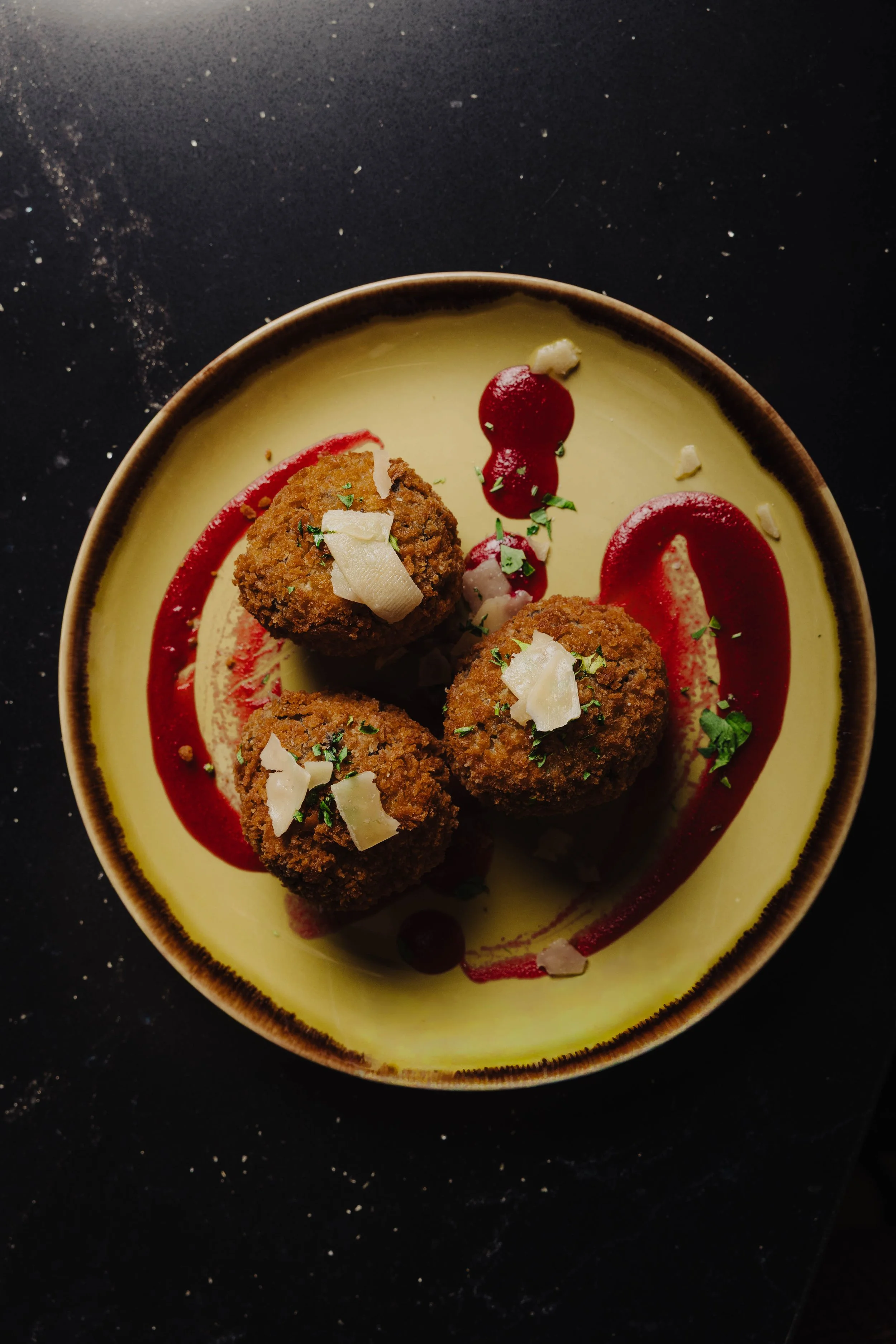 Fried appetizers garnished with shredded cheese and herbs, served with a streak of red sauce and a drizzle of yellow sauce on a yellow plate.